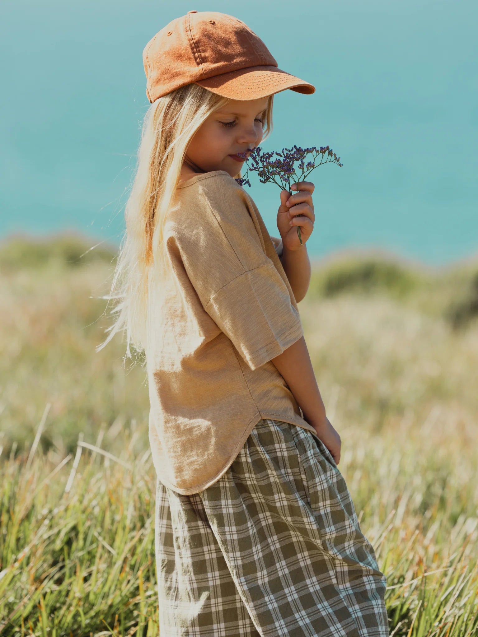 A young child with long blonde hair stands in a grassy field by the seaside, holding and sniffing a small bouquet of purple flowers. The child is wearing an olive green print shirt, a rust-colored cap, a unisex design plaid skirt, and VALENCIA BYRON BAY's Rio Pant Olive, with the serene blue ocean in the background.