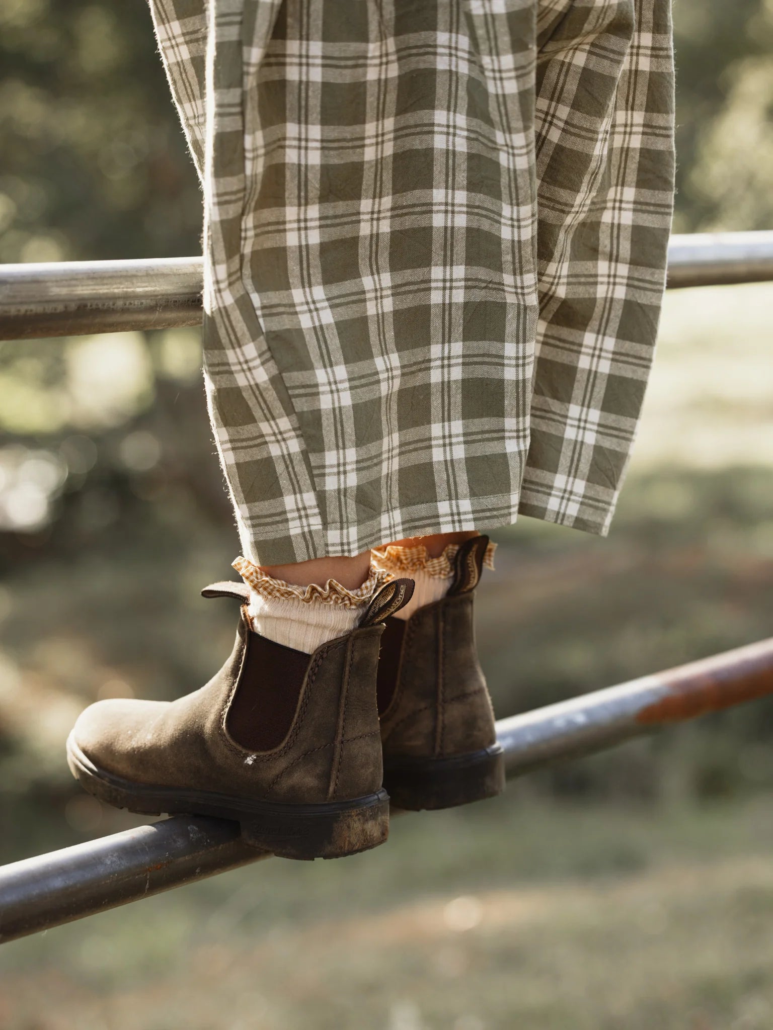 A person wearing brown suede boots, matching cream-colored frilly socks, and the Rio Pant Olive from VALENCIA BYRON BAY is balancing on a metal fence. The scene is set outdoors with blurred greenery in the background, suggesting a natural, serene environment.