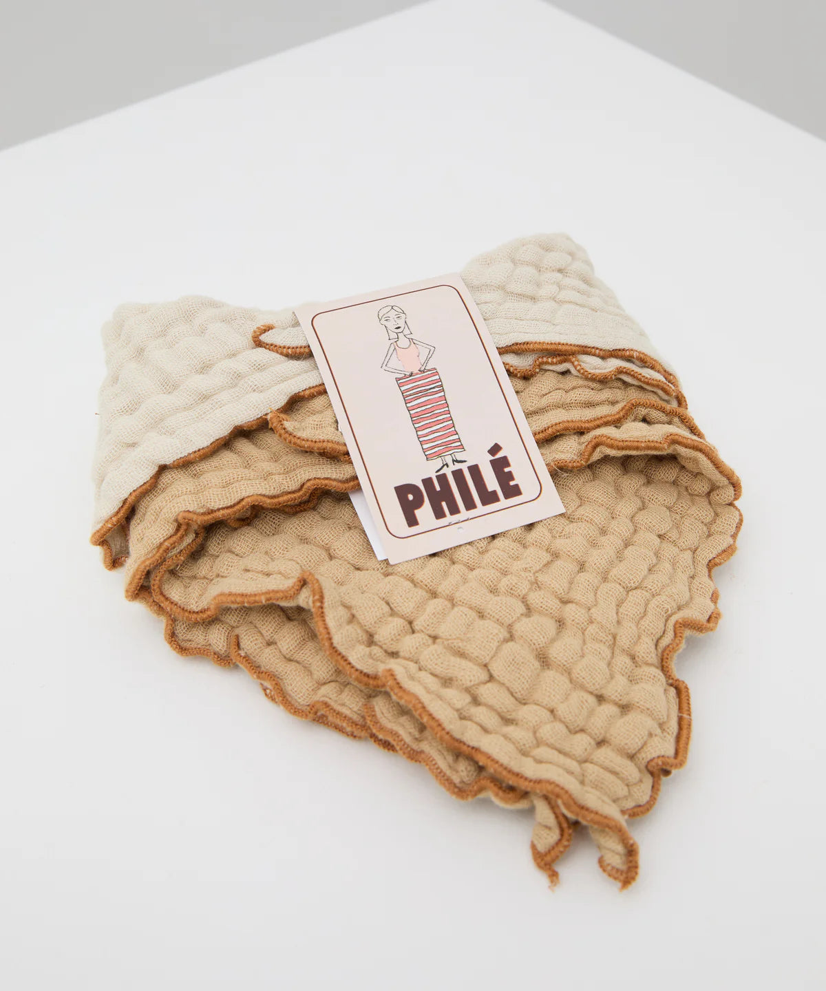 A stack of cacao-colored Philé muslin cotton bibs by PHILÉ is neatly folded in a triangular bandana style. On top, a card shows an illustrated woman and "PHILÉ" in bold letters. This is the Cotton Bib Double Pack Cacao set.