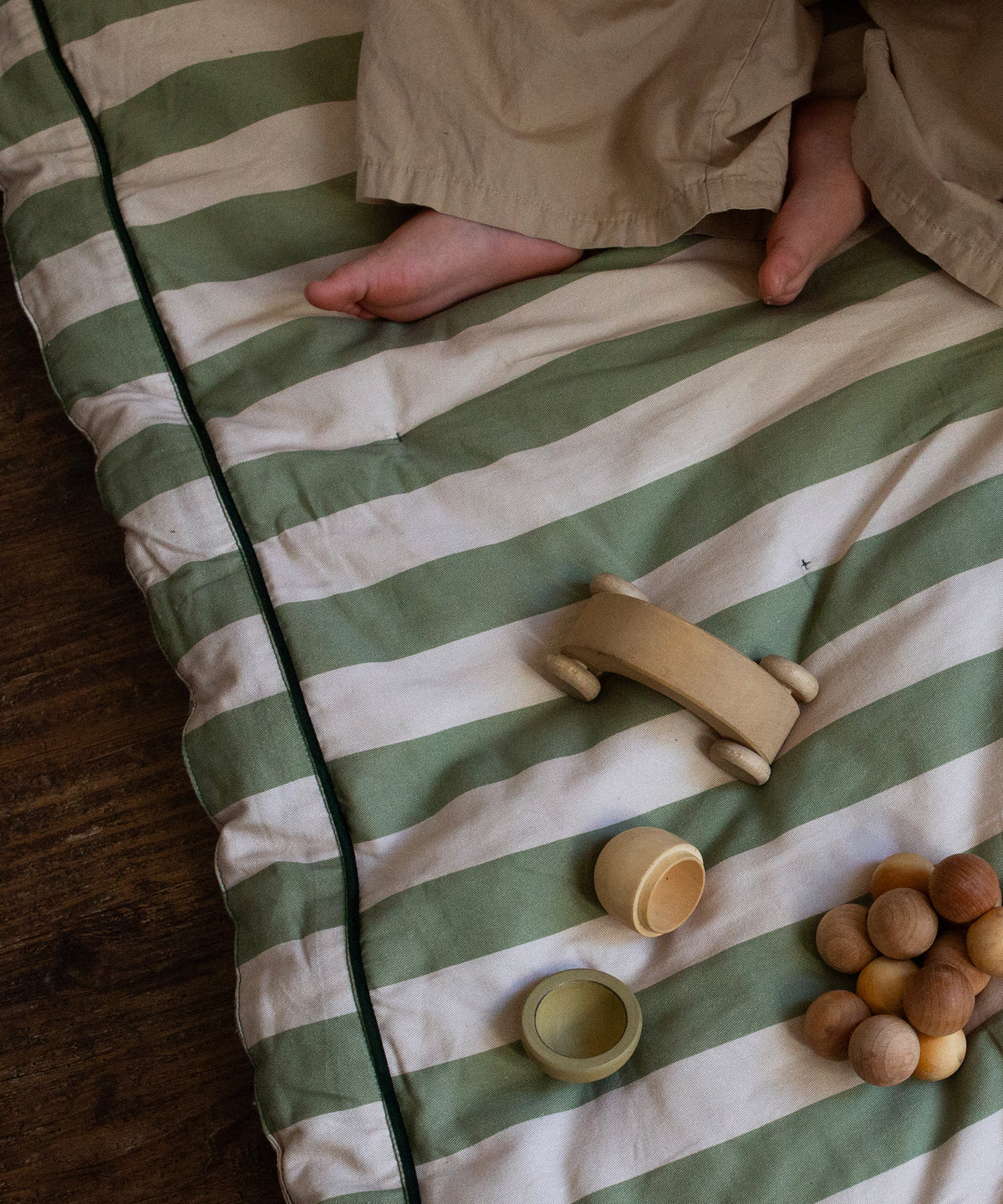 A child’s bare feet and beige pants rest on the Philé - Striped Play Mat Green by PHILÉ, with wooden toys scattered nearby.