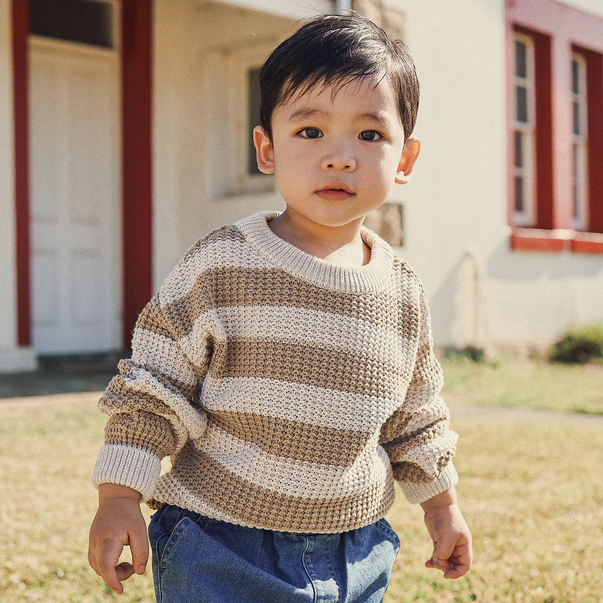 A young child with short dark hair, wearing the KYND BABY Organic Textured Knit Jumper in Caramel Stripe and blue jeans, stands on grass outside a white building with red trim.