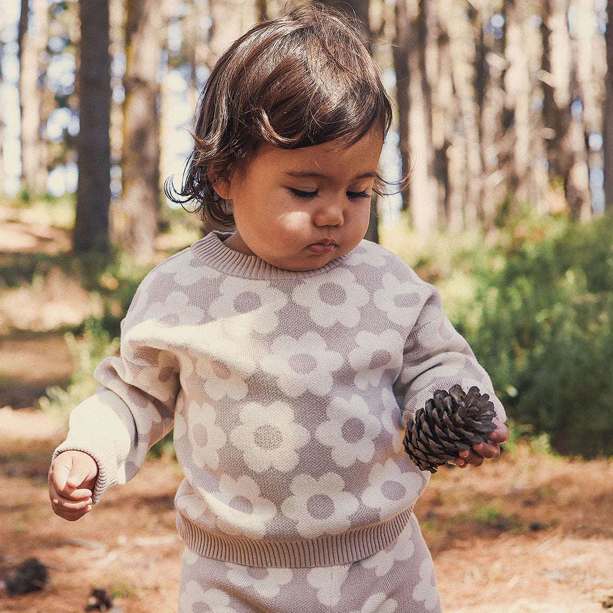A toddler in the KYND BABY Kynd Baby - Knit Jumper Neutral Flower, featuring white flower patterns, stands in a sunlit forest, looking down while holding a pinecone.
