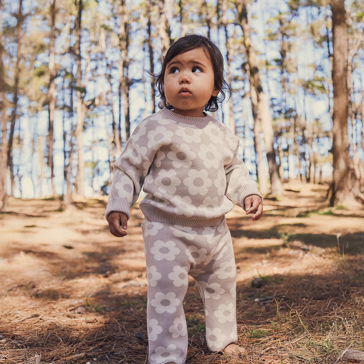 A toddler stands barefoot on a sunlit forest floor, wearing KYND BABY’s Kynd Baby - Knit Flare Pants Neutral Flower, featuring large floral patterns in light-colored organic cotton with a matching top.