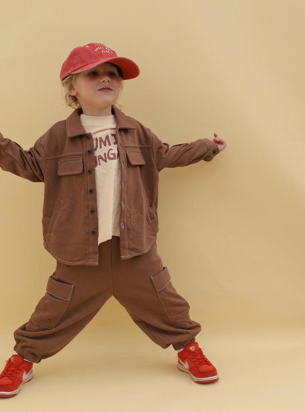A young child stands against a beige background, wearing SMALL SWIM CLUB's Choccy Cargo Trackies with a brown jacket, white printed t-shirt, red cap, and red sneakers. Their expressive pose with outstretched arms highlights playful spirit.