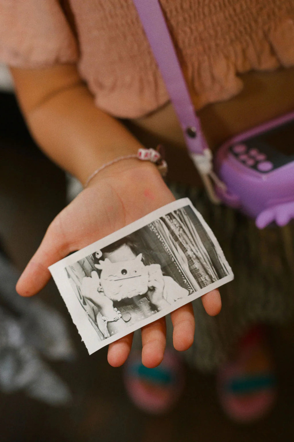A child holds a black-and-white instant photo printed by the OH MY FRANKIE Oh My Snap Instant Print Camera Pink, wearing a pink textured top, colorful beaded bracelet, and carrying a purple camera case over their shoulder.