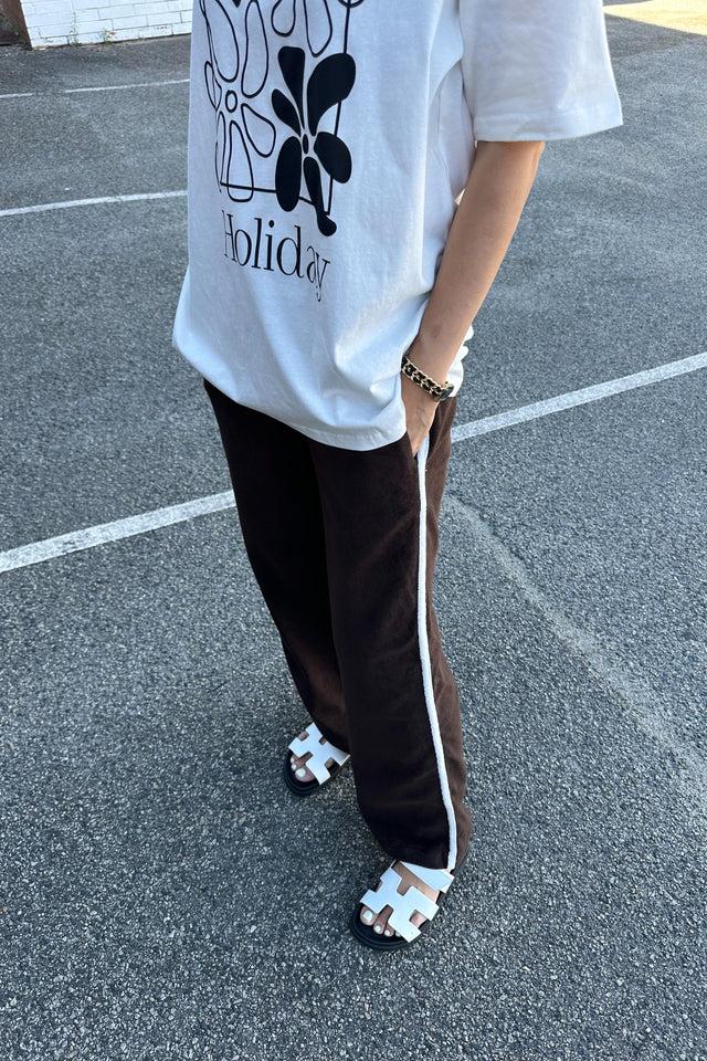 A person stands on a pavement wearing a white T-shirt with a floral design and the word "Holiday" printed on it, black Piper Racer Pants Dark Chocolate by TINY TROVE featuring relaxed legs and an elasticated waistband, and white slip-on sandals. They have a gold chain bracelet on their left wrist, and their right hand is casually in their pocket.