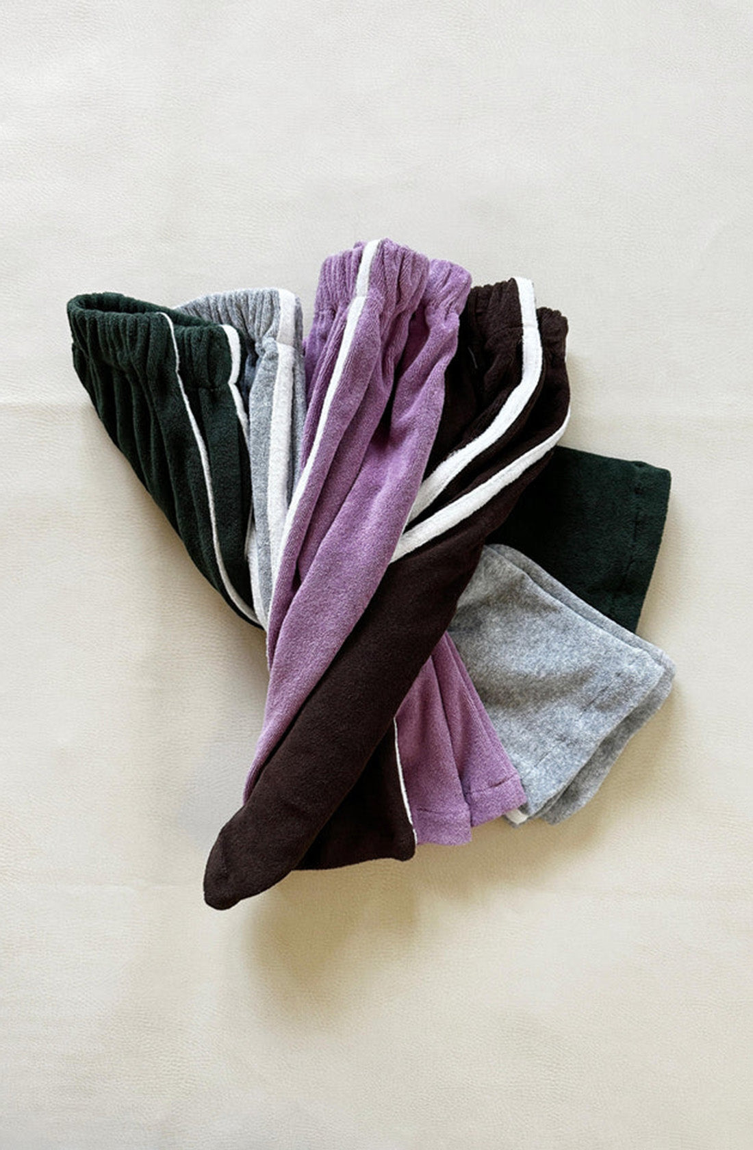 A collection of Darcy Racer Pants Wisteria from TINY TROVE, in a variety of colors such as dark green, light gray, purple, dark brown, and dark gray, is displayed on a light beige surface. Made from premium terry towelling with a comfortable elastic waistband, the pants are neatly stacked with their waists aligned.