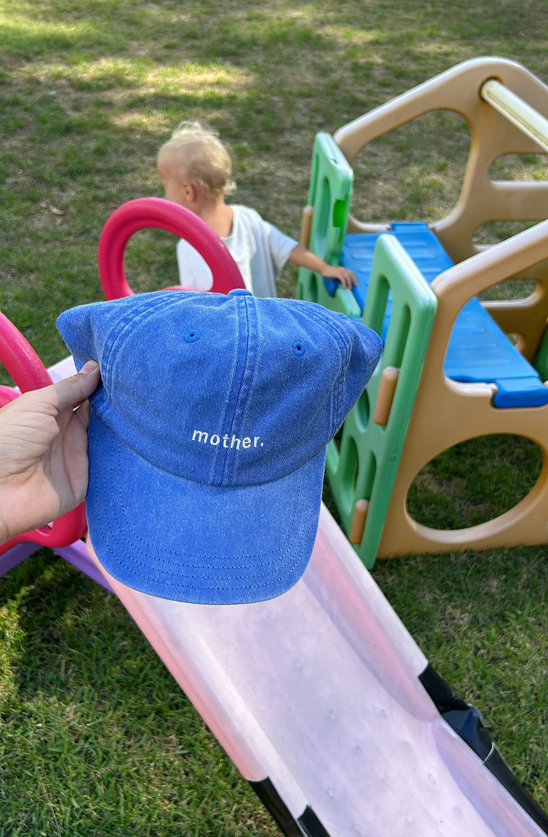 A person holding a blue cap with the word mother on it, in the background it a kid playing on play equipment.