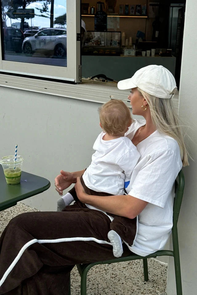 A woman in a white cap and white t-shirt with an elasticated waistband, paired with Piper Racer Pants in Dark Chocolate by TINY TROVE, is sitting at an outdoor café holding a baby on her lap. A plastic cup with a green drink and a striped straw sits on the table in front of her. The café has an open window, reflecting cars in the glass.