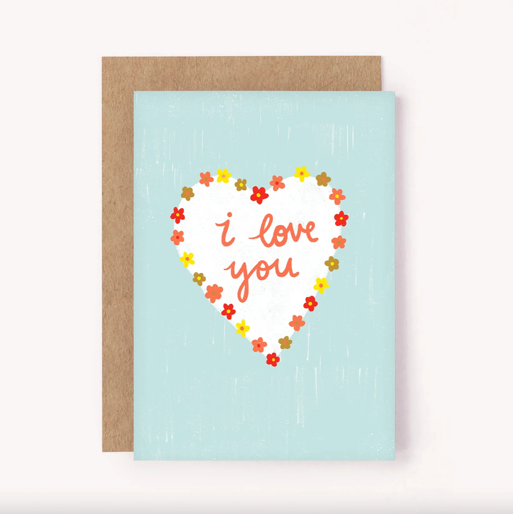 The LAUREN SISSONS STUDIO Greeting Card 'I Love You' features a light blue background with a white heart in the center, surrounded by small red, yellow, and orange flowers. The text "I love you" is elegantly written in cursive red letters inside the heart. Crafted on recycled paper, this charming card comes with a brown envelope.