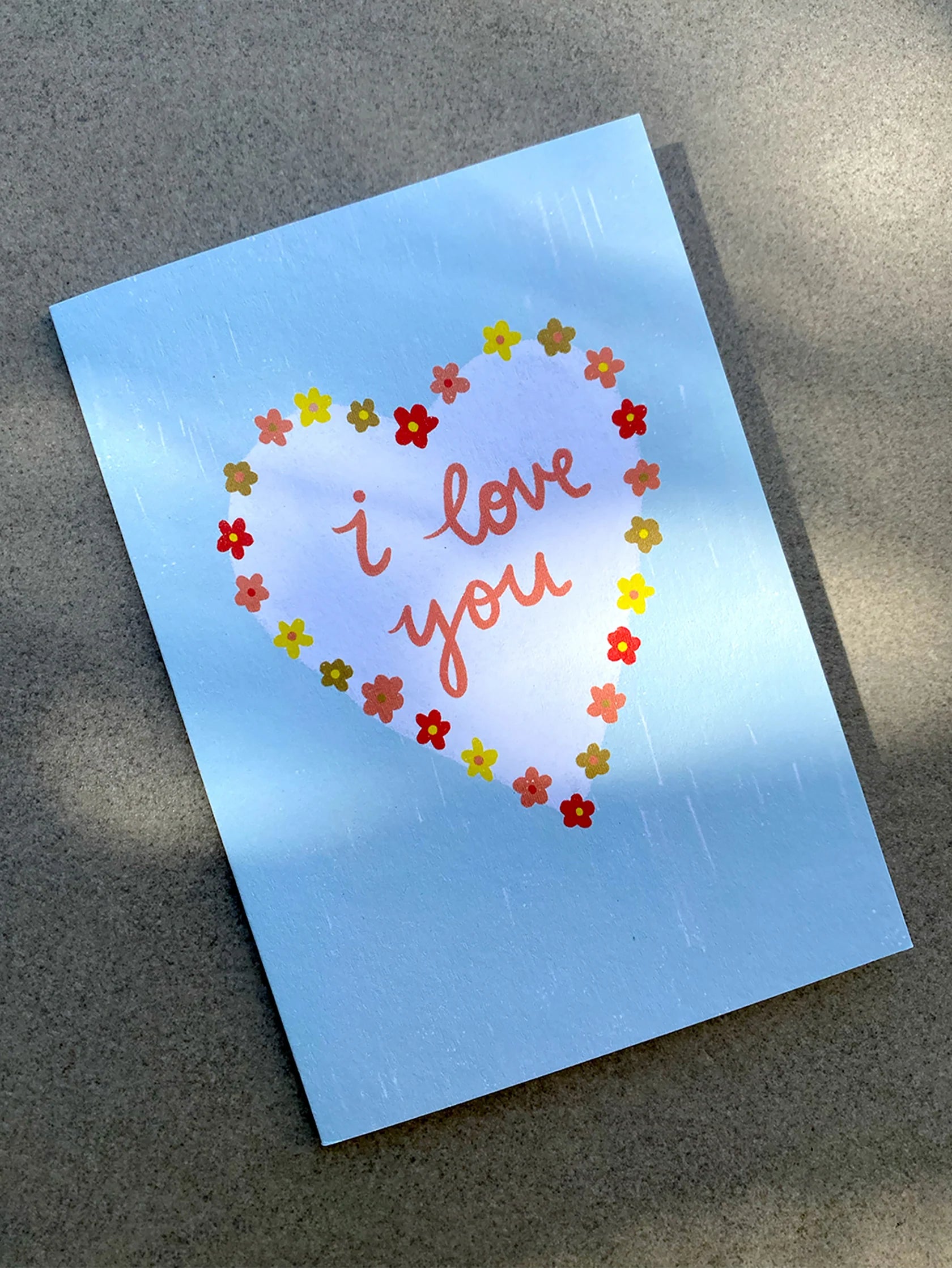 A light blue illustrated greeting card from LAUREN SISSONS STUDIO rests on a gray surface. The 'I Love You' card showcases a heart-shaped wreath made of small, colorful flowers. Inside the heart, in pink cursive text, it reads "I love you." Crafted from recycled paper and bathed in dappled sunlight, this card epitomizes sustainable production.