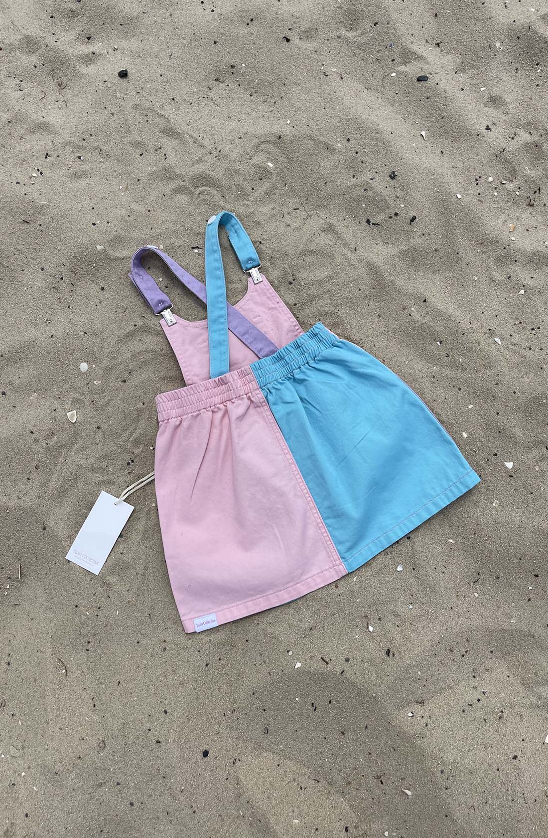 A color-blocked pink and blue children's dress, the "Bowie Dress Colourblock" by TWIN COLLECTIVE, made from sustainable 100% cotton with adjustable rainbow-colored straps lies on sandy ground. A white tag is attached, indicating it is brand new.