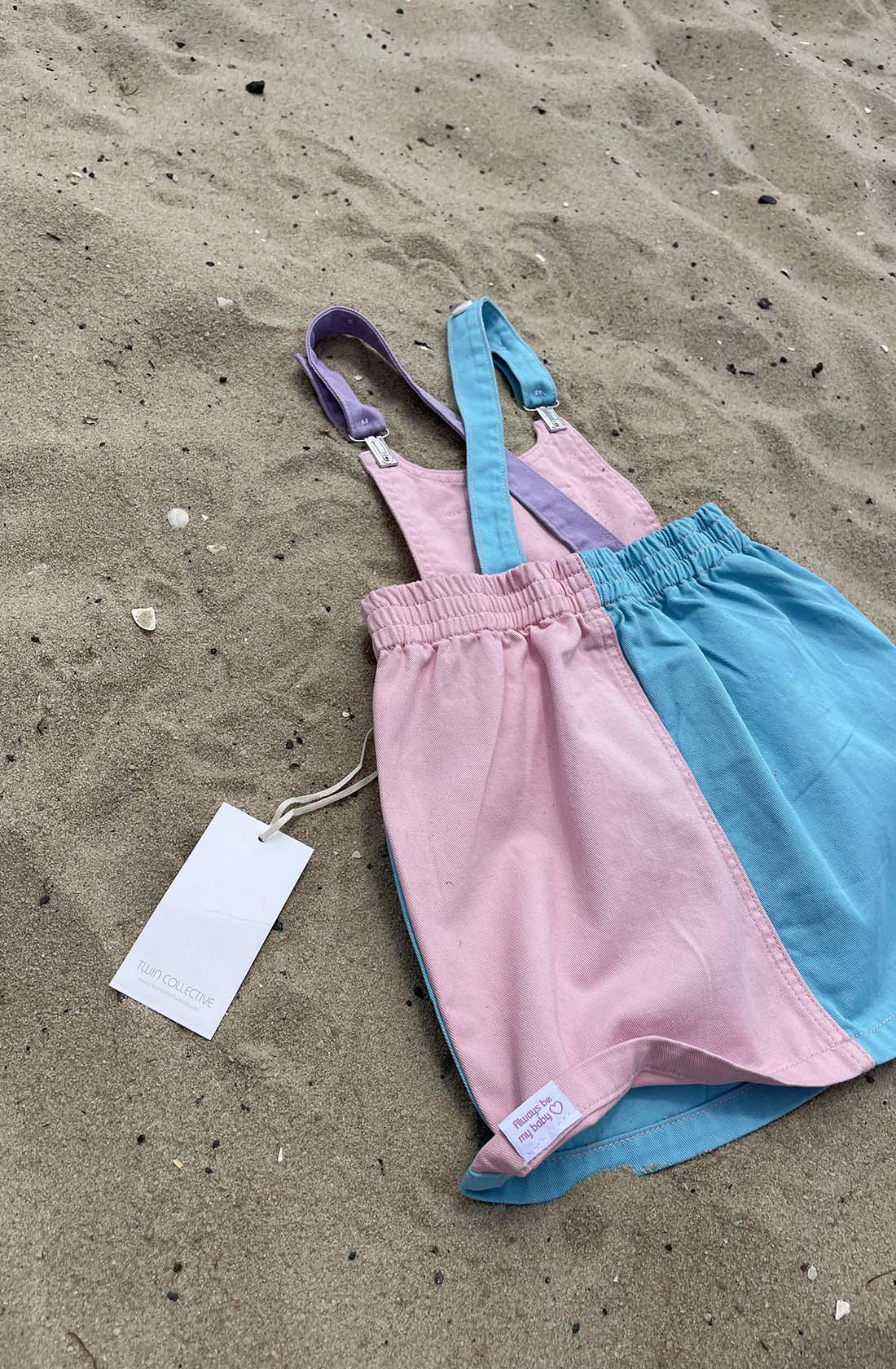 The Bowie Dress Colourblock by TWIN COLLECTIVE, a pink and blue children's romper made from sustainable 100% cotton, is laid out on sandy ground with small shells and pebbles scattered around. The romper features lilac straps, an elastic waistband, and a clothing tag attached.