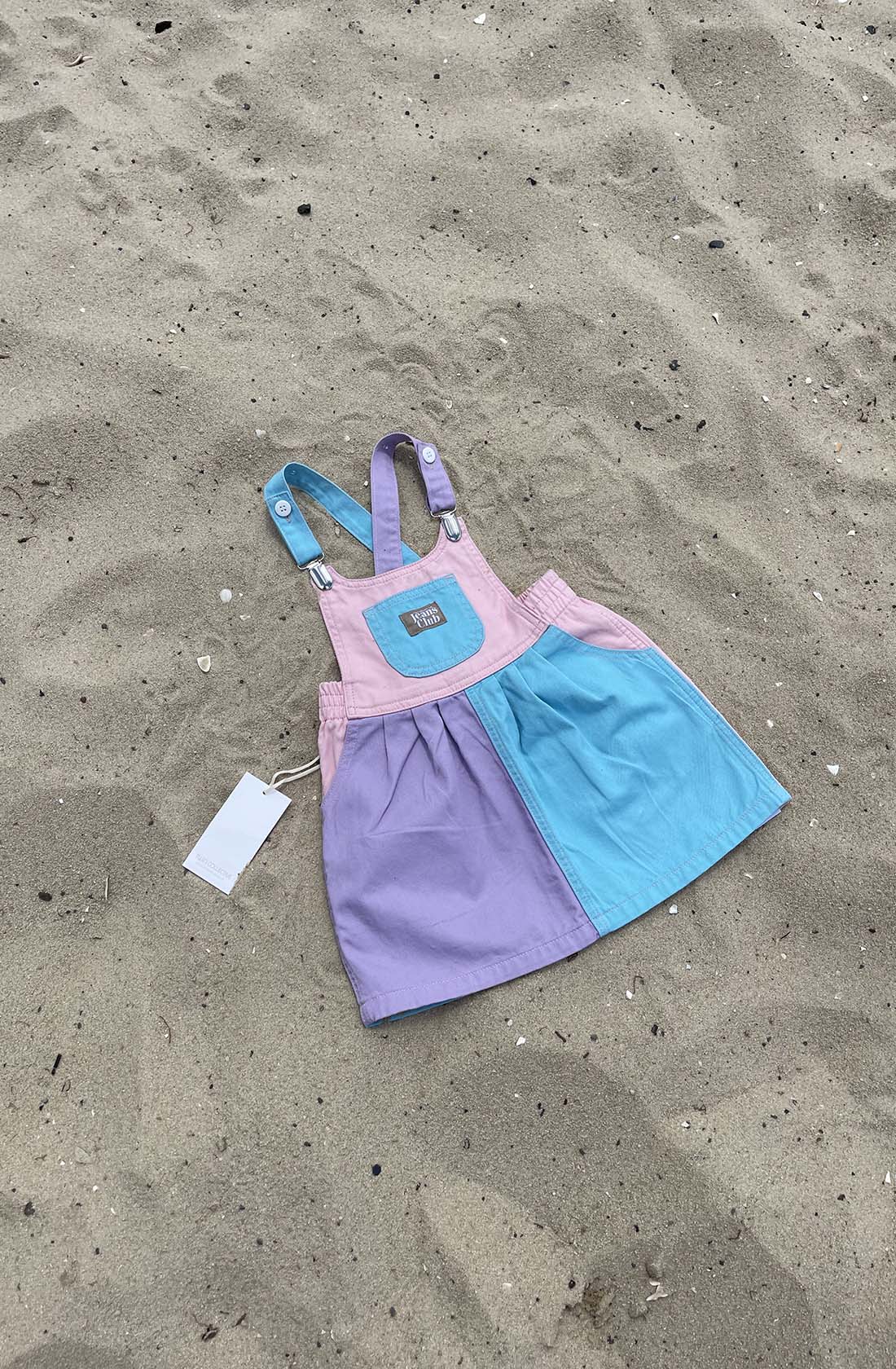 A small, colorful Bowie Dress Colourblock from TWIN COLLECTIVE, featuring panels in pink, blue, and purple, is lying on sandy ground. This sustainable dress made of 100% cotton includes two shoulder straps with metal clasps and a front pocket adorned with a label. A white tag is attached to the dress.