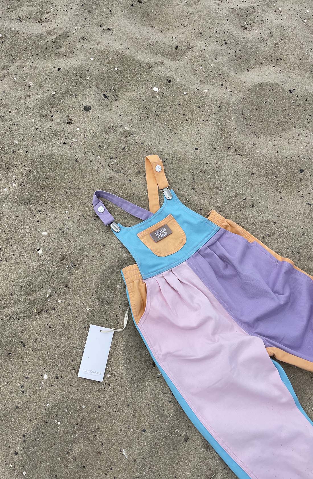 The Bowie Bubble Colourblock overall for children from TWIN COLLECTIVE, featuring sustainable cotton in pastel blocks of purple, pink, blue, and orange, lies on the sandy ground. These kids' denim overalls include metal clasps on the adjustable straps and a tag attached to one side.