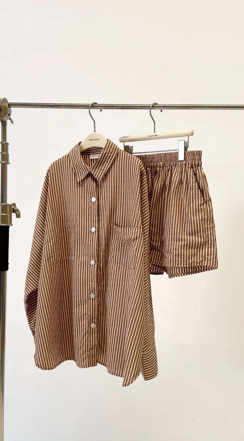The TINY TROVE Women's Isla Stripe Cotton Set in Caramel/White Stripe, featuring a relaxed fit oversized shirt and elastic waistband shorts, hangs on a metal rack against a plain white background.