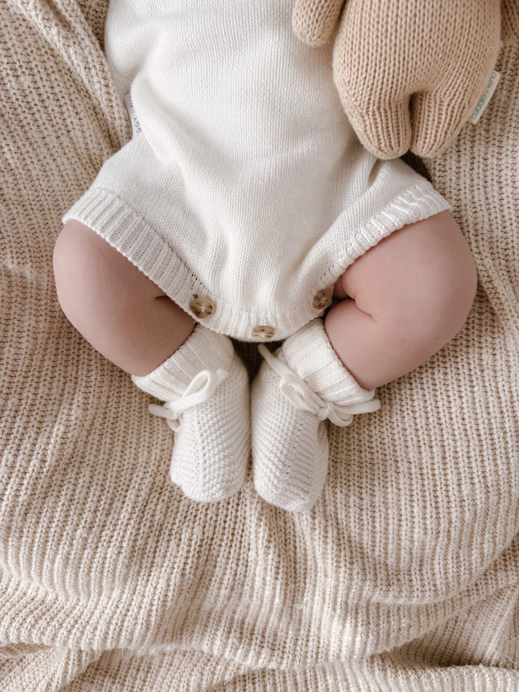 A baby in a cream knit onesie sits on a beige blanket, wearing ZIGGY LOU Booties Milk. The focus is on the baby's legs and feet, with their hands resting beside them.