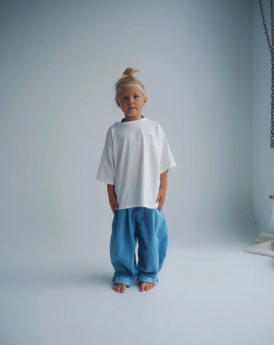 A young child with blonde hair in a topknot stands barefoot in a minimalist studio, wearing an oversized white T-shirt and ZIGGY ZAZA's Ziggy Zaza - Suki Denim Jean Light Wash Denim, looking directly at the camera.