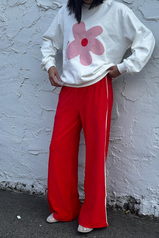 Standing against a textured white wall, the individual wears a TINY TROVE white sweatshirt with a pink flower design, paired with Piper Racer Pants Red that feature an elasticated waistband and white stripes. Their ensemble is completed with red and white shoes, hands confidently resting on their hips.