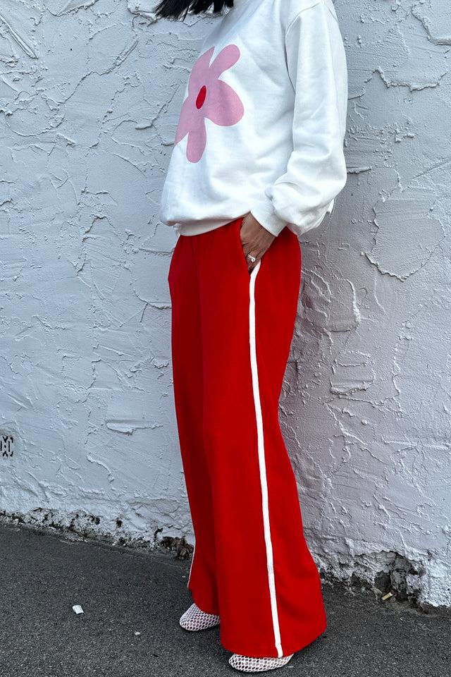 A person stands against a textured white wall wearing a white sweater with a pink flower design, TINY TROVE's Piper Racer Pants Red featuring an elasticated waistband and white side stripes, and white mesh shoes. Their hands are in their pockets, and their head is not visible.