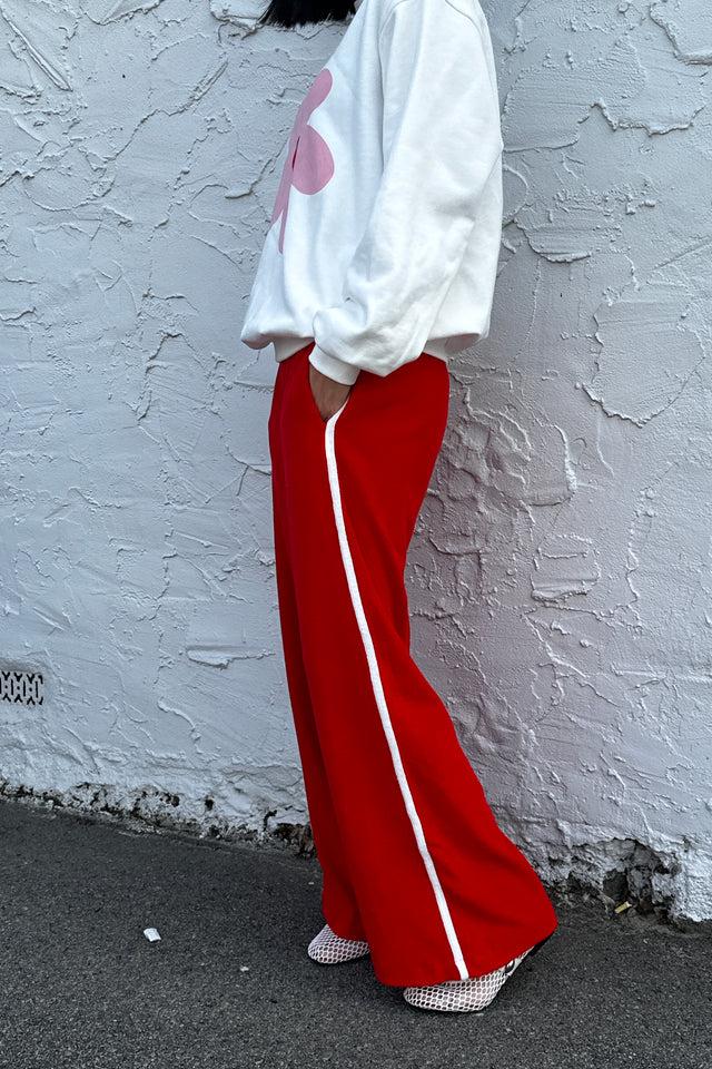 A person standing against a textured wall is wearing a white sweatshirt with a pink heart, TINY TROVE Piper Racer Pants in red featuring an elasticated waistband and mid-waisted fit, and checkered shoes.