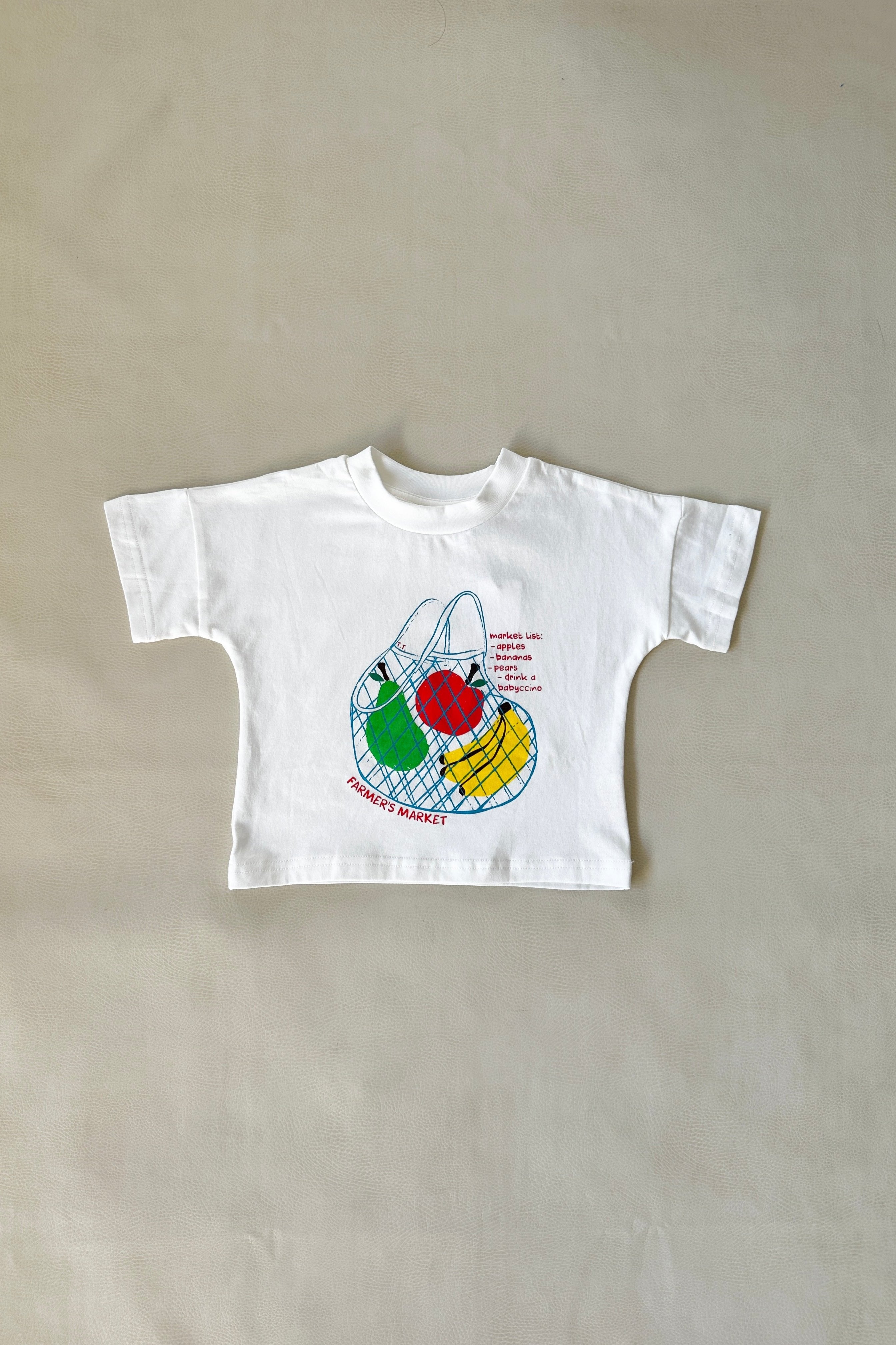 A toddler-sized Farmer's Market Relaxed Tee in white, designed by TINY TROVE, features a vibrant pattern with a mesh bag filled with red, yellow, and green fruits and the text "Farmer's Market" on a plain light background.