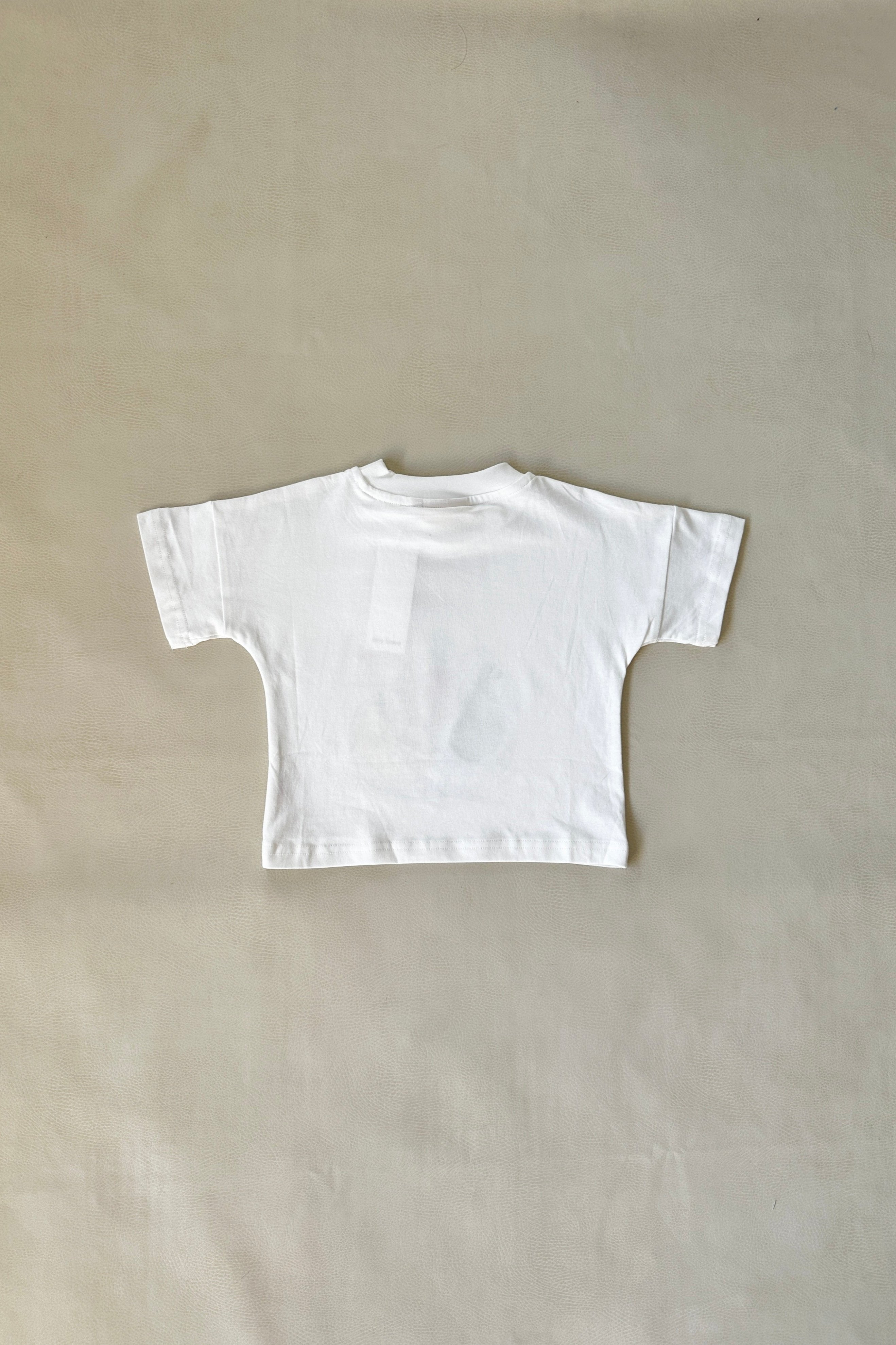 The Farmer's Market Relaxed Tee White by TINY TROVE is displayed laid flat on a light beige background, highlighting the back with short sleeves and a round neckline.