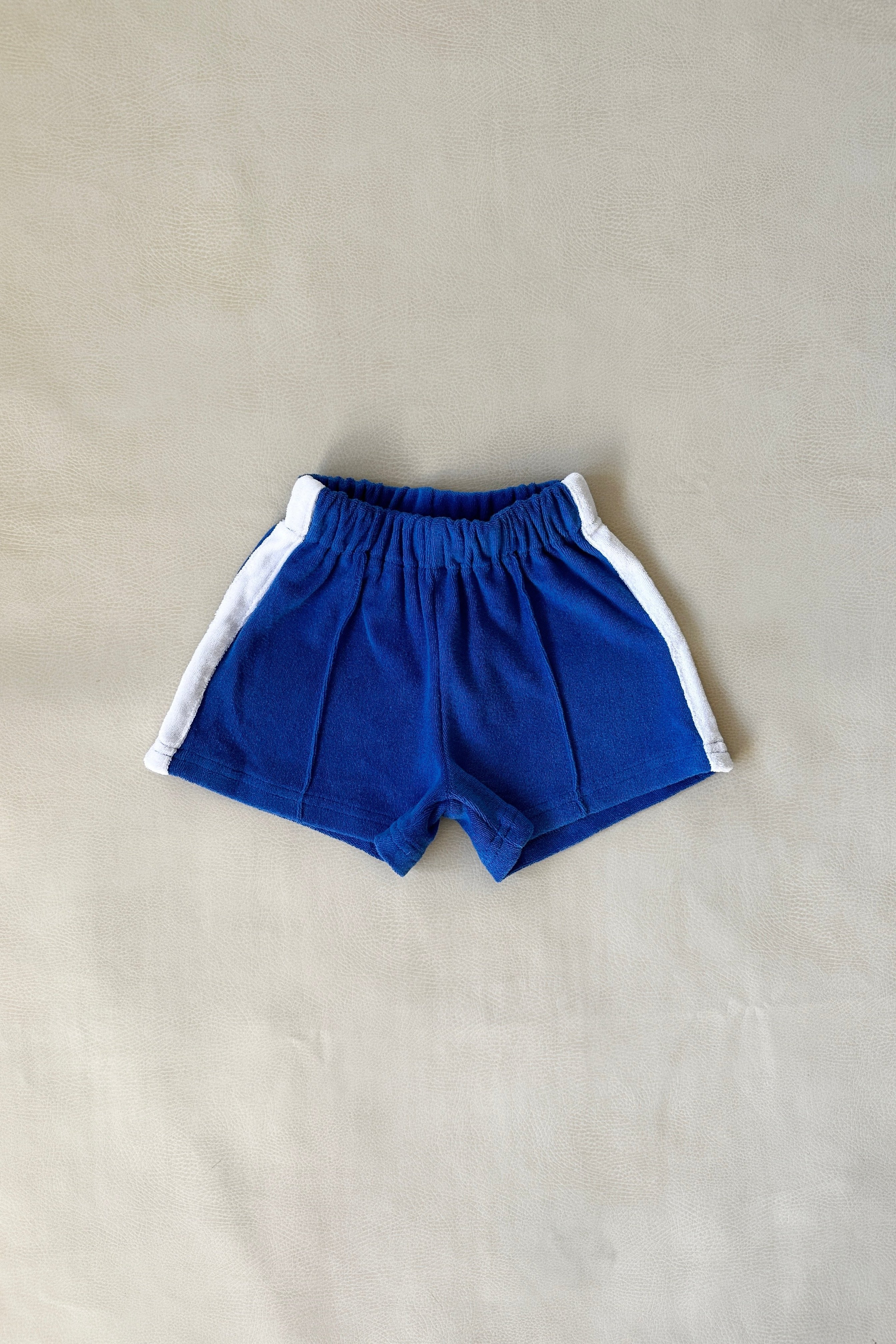 The Atlas Cotton Terry Towelling Set in Royal Blue by TINY TROVE, size 6, features blue children's shorts with white stripes on the sides. Made from soft terry cotton, it has a relaxed fit and an elastic waistband for comfort. Displayed flat on a beige surface.