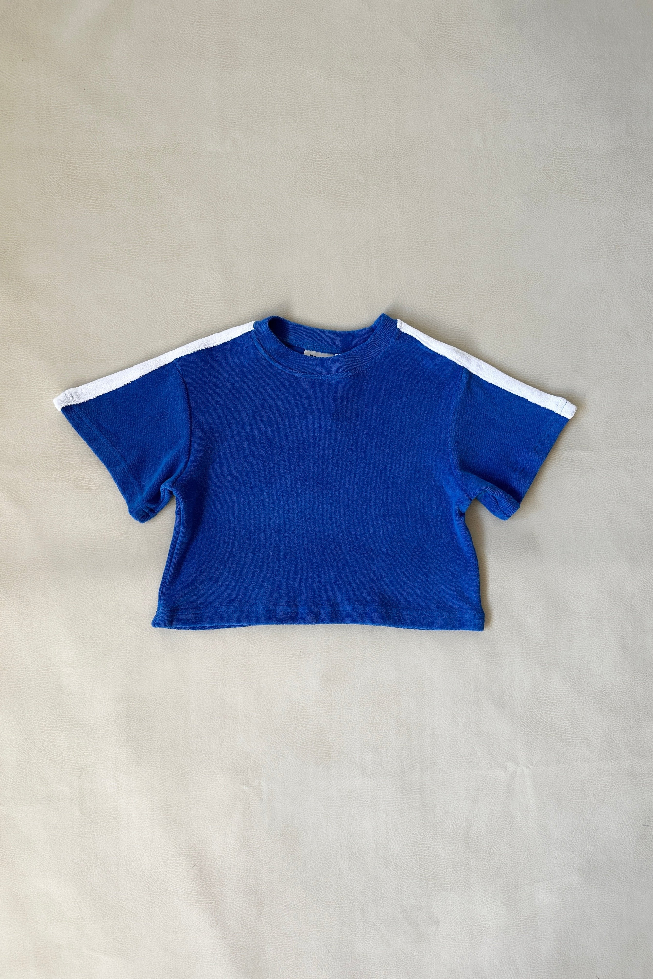 A royal blue short-sleeve crop top from TINY TROVE's Atlas Cotton Terry Towelling Set, Size 6, featuring white sleeve stripes, is displayed on a plain light backdrop.