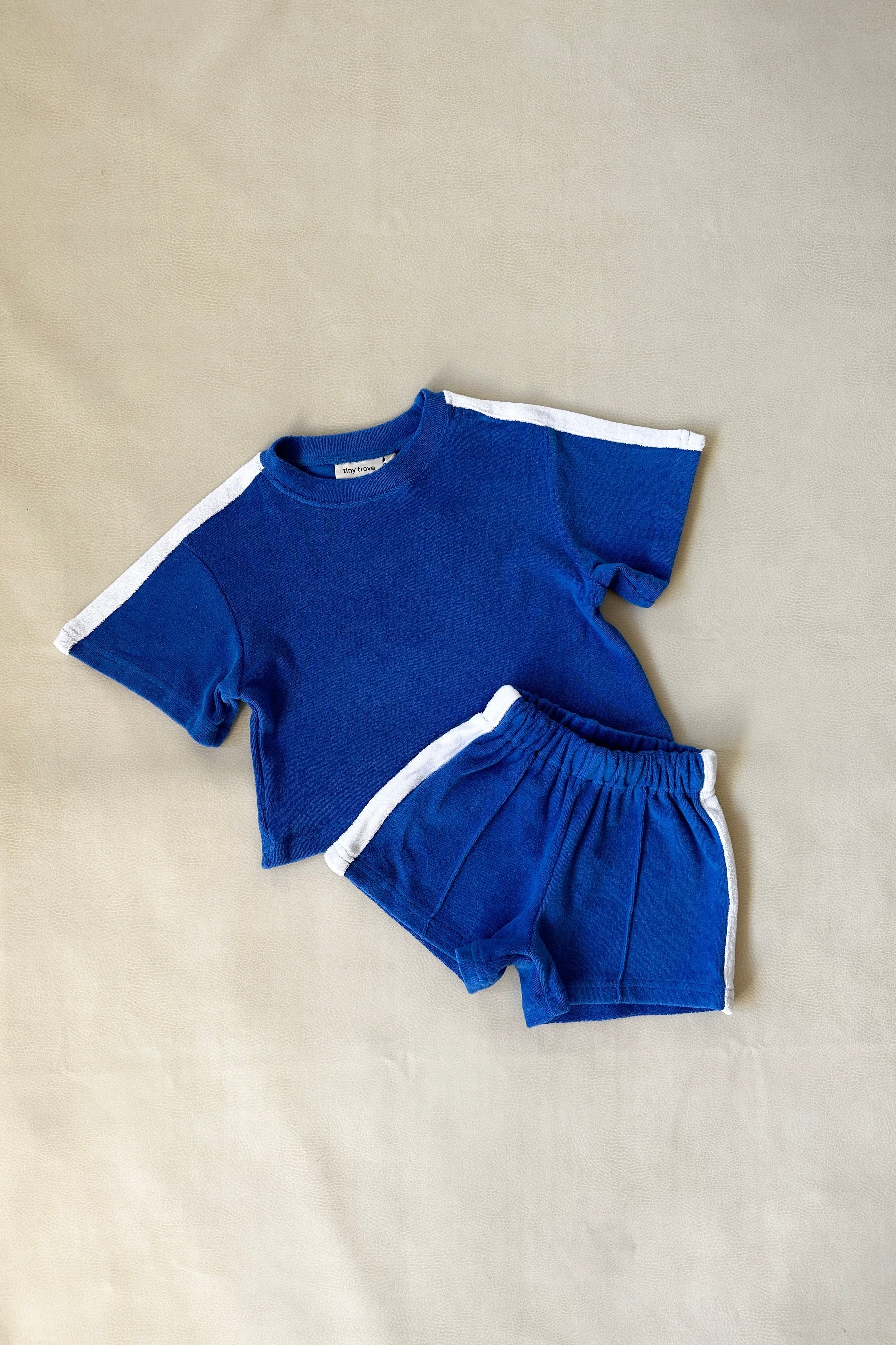 The TINY TROVE Atlas Cotton Terry Towelling Set in Royal Blue, featuring a relaxed-fit toddler tee and shorts with white contrast trim, is laid flat on a beige background.