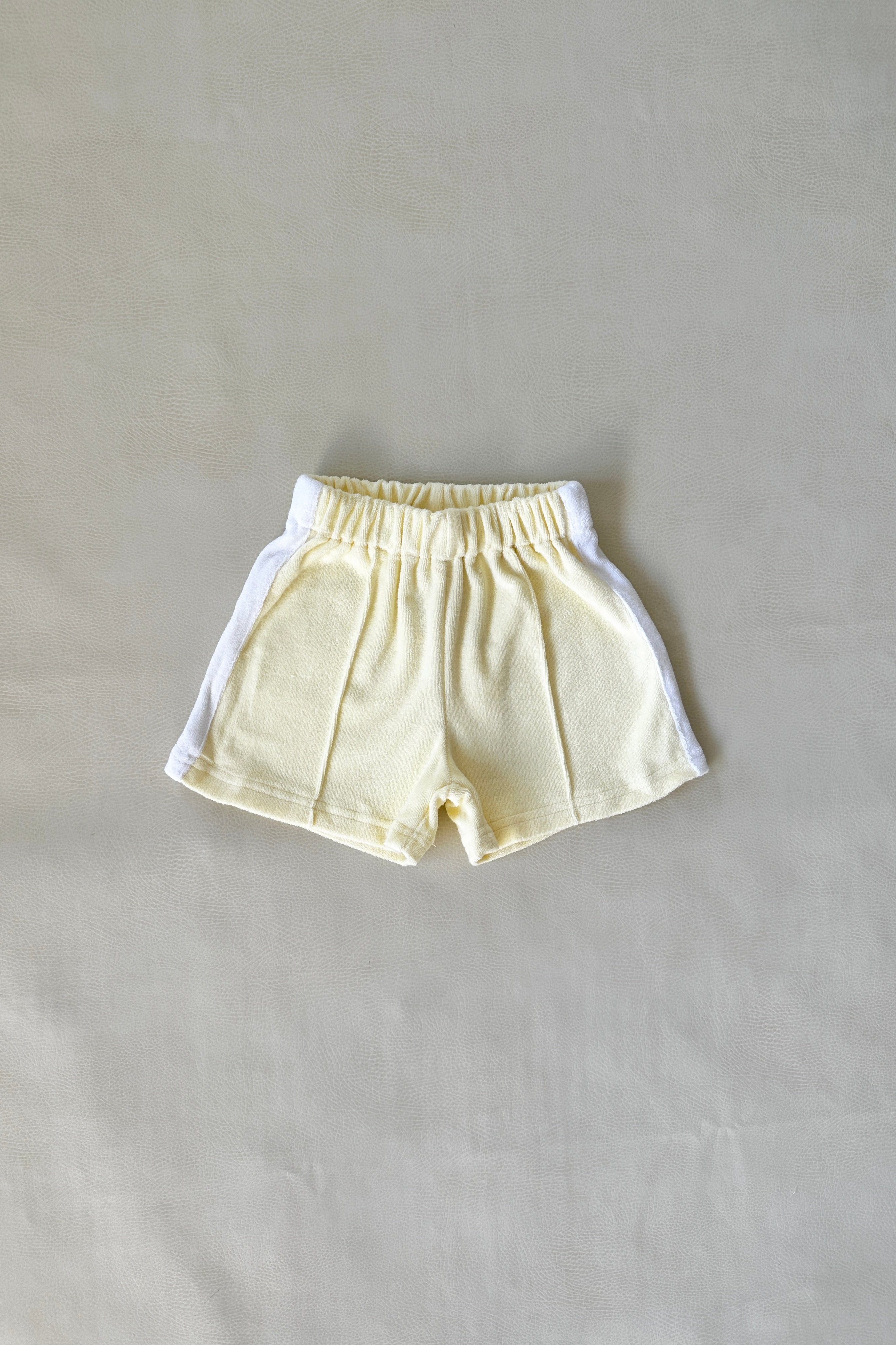 A piece from the TINY TROVE collection, the Atlas Cotton Terry Towelling Set in Lemon Drop showcases a pair of light yellow toddler shorts with white trim, elegantly presented against a neutral backdrop.