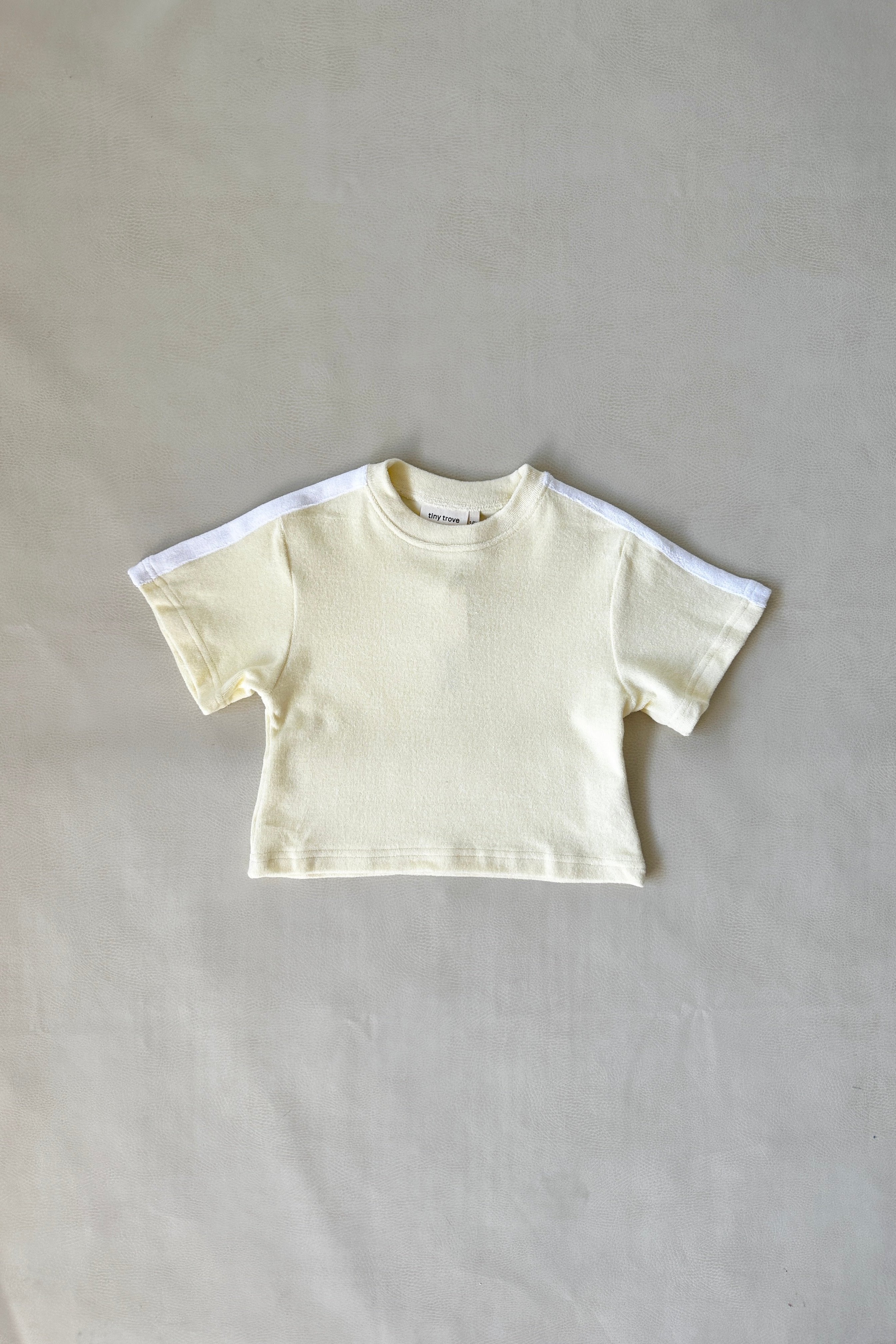 The Atlas Cotton Terry Towelling Set - Lemon Drop by TINY TROVE lies flat on a plain, light gray surface. The small, cream-colored infant T-shirt is made from soft terry towelling cotton and features a relaxed fit with short sleeves and a simple design.