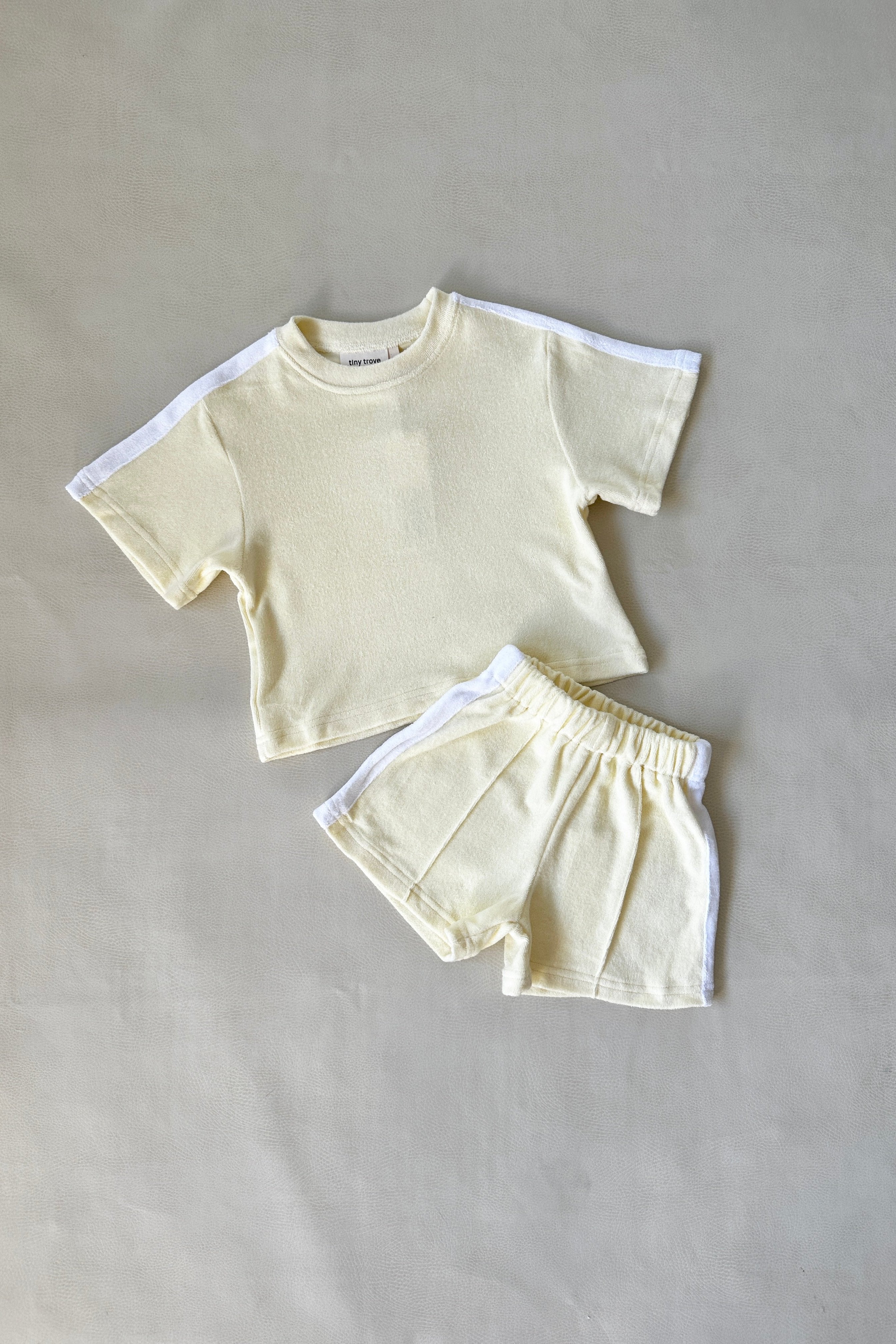 Introducing the TINY TROVE Atlas Cotton Terry Towelling Set in Lemon Drop—a delightful light yellow toddler outfit. This set includes a relaxed-fit tee and shorts, crafted from soft terry towelling cotton. Both pieces are adorned with white side stripes, and the shorts feature an elastic waistband for added comfort. Presented on a neutral background, this ensemble effortlessly showcases its cozy style.