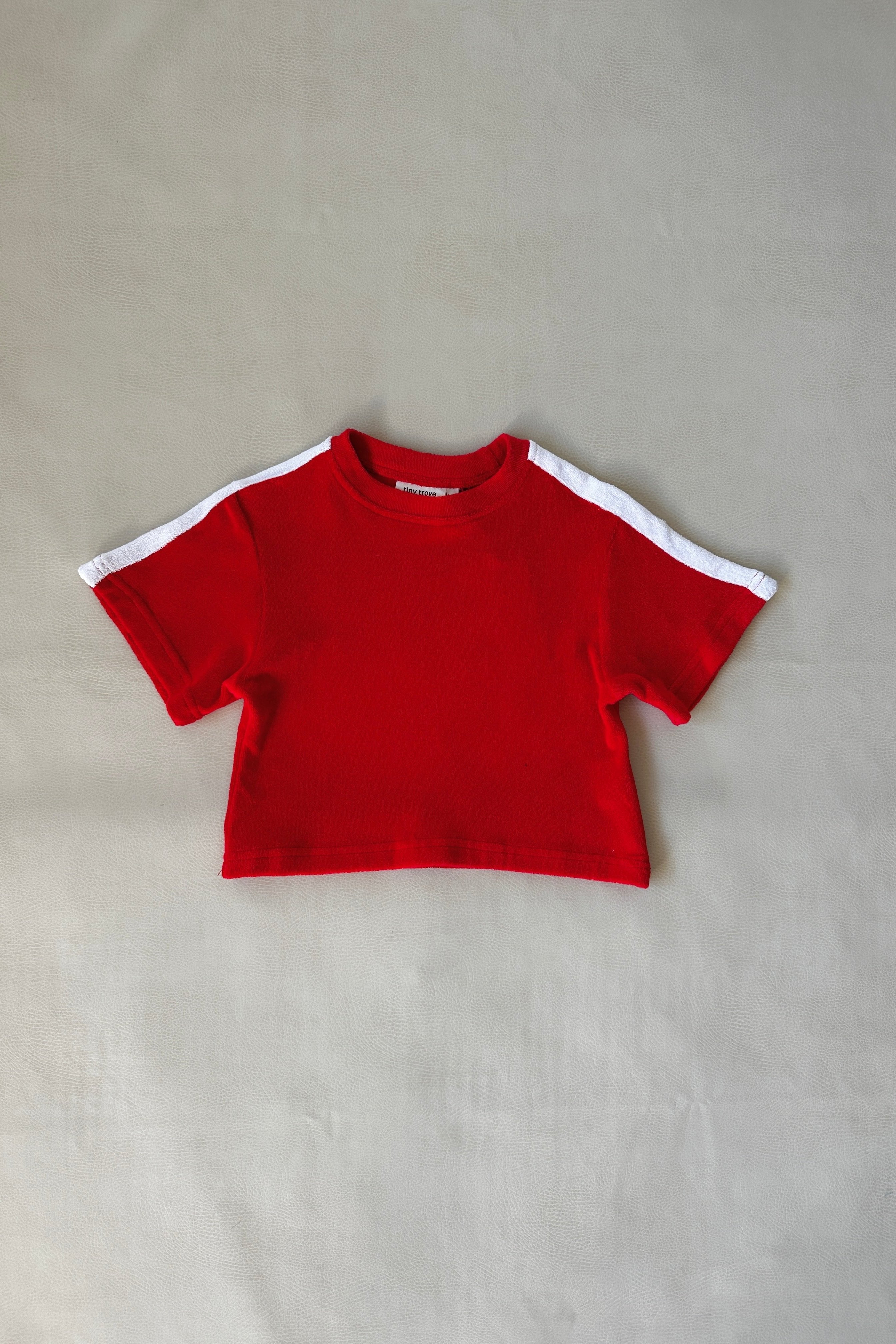 The Atlas Cotton Terry Towelling Set in Red by TINY TROVE is displayed flat on a simple beige backdrop, showcasing the white contrast along the shoulders and sleeves that pairs perfectly with any casual tee and shorts outfit.