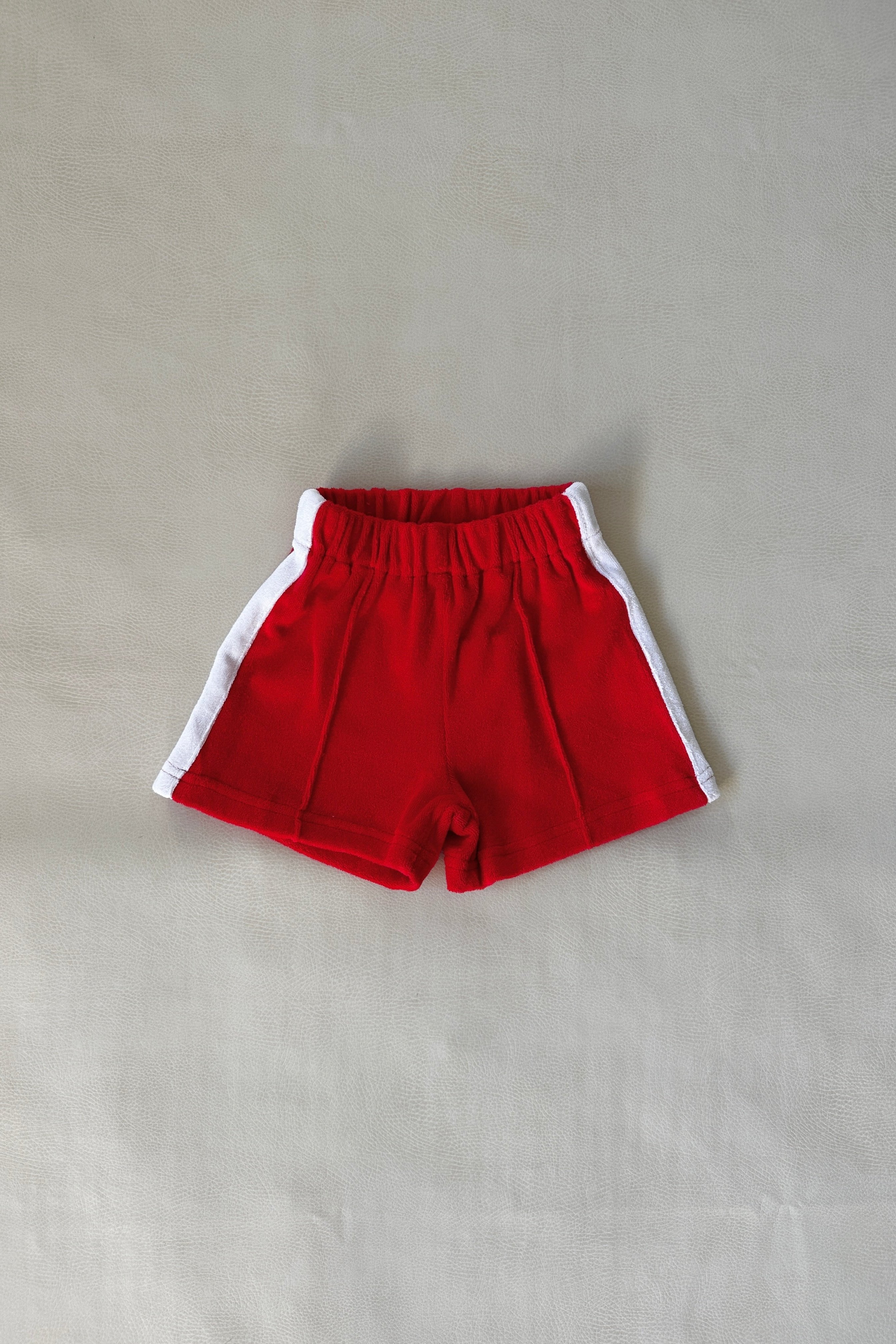 The Atlas Cotton Terry Towelling Set Red by TINY TROVE includes children's shorts in a relaxed fit, adorned with white contrast side stripes. These red shorts are displayed lying flat on a neutral background and feature an elastic waistband, perfectly designed for casual wear.