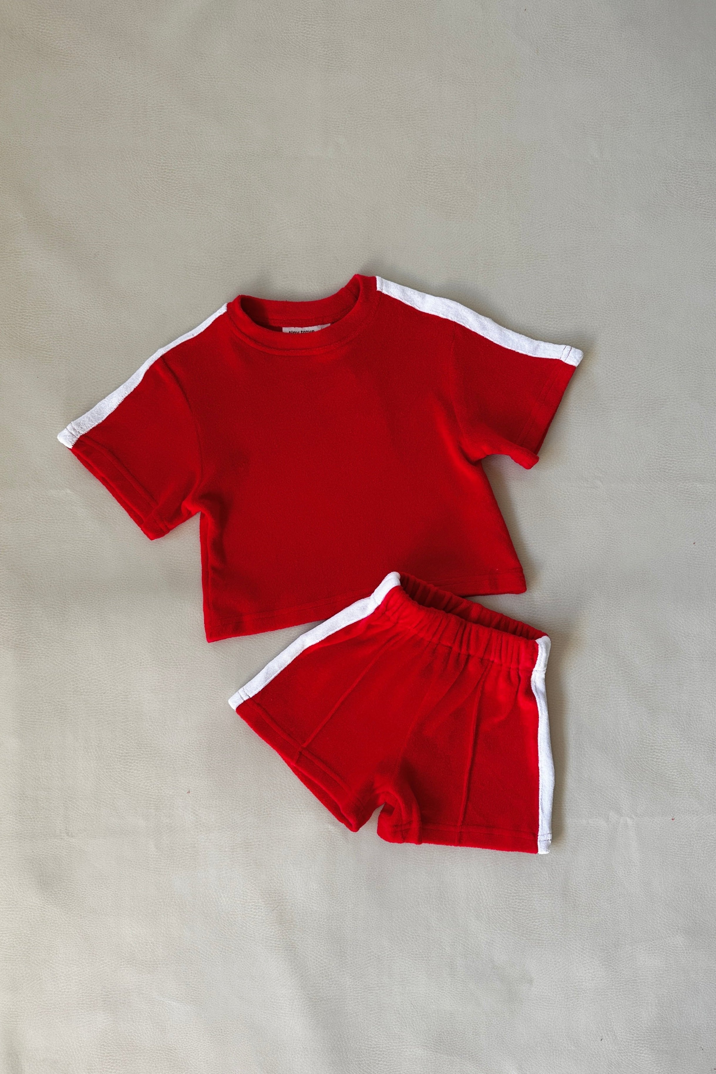 The Atlas Cotton Terry Towelling Set Red by TINY TROVE is a charming children's athletic outfit featuring a relaxed fit and white contrast stripes. This set includes a short-sleeved crop top and matching shorts, displayed on a light-colored background.