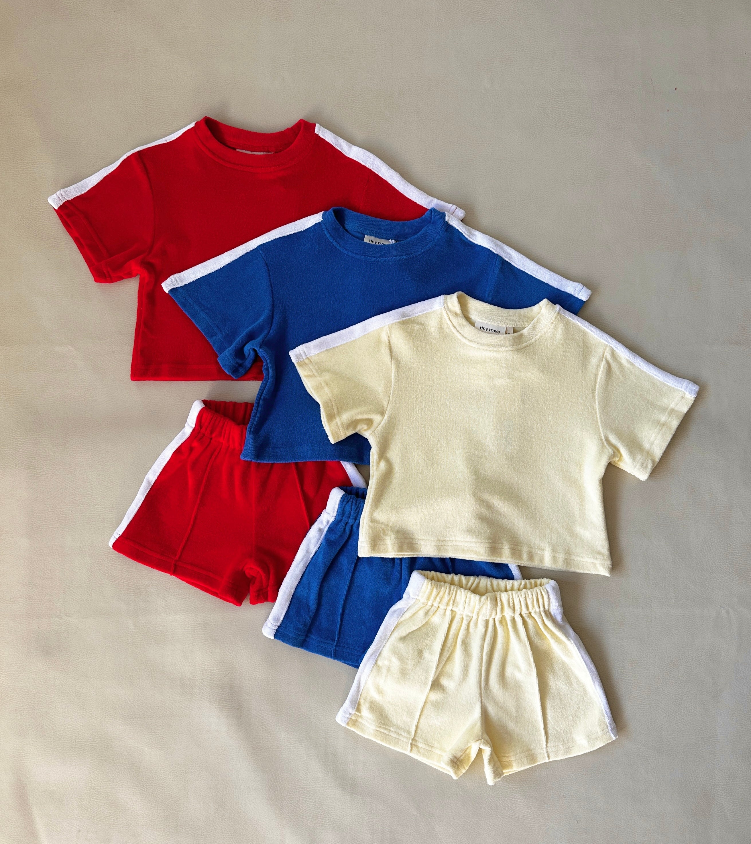 A trio of Atlas Cotton Terry Towelling Sets by TINY TROVE is displayed on a flat surface, featuring a relaxed fit in red, blue, and lemon drop. Each set includes white stripes accenting the sleeves and sides of the shorts.