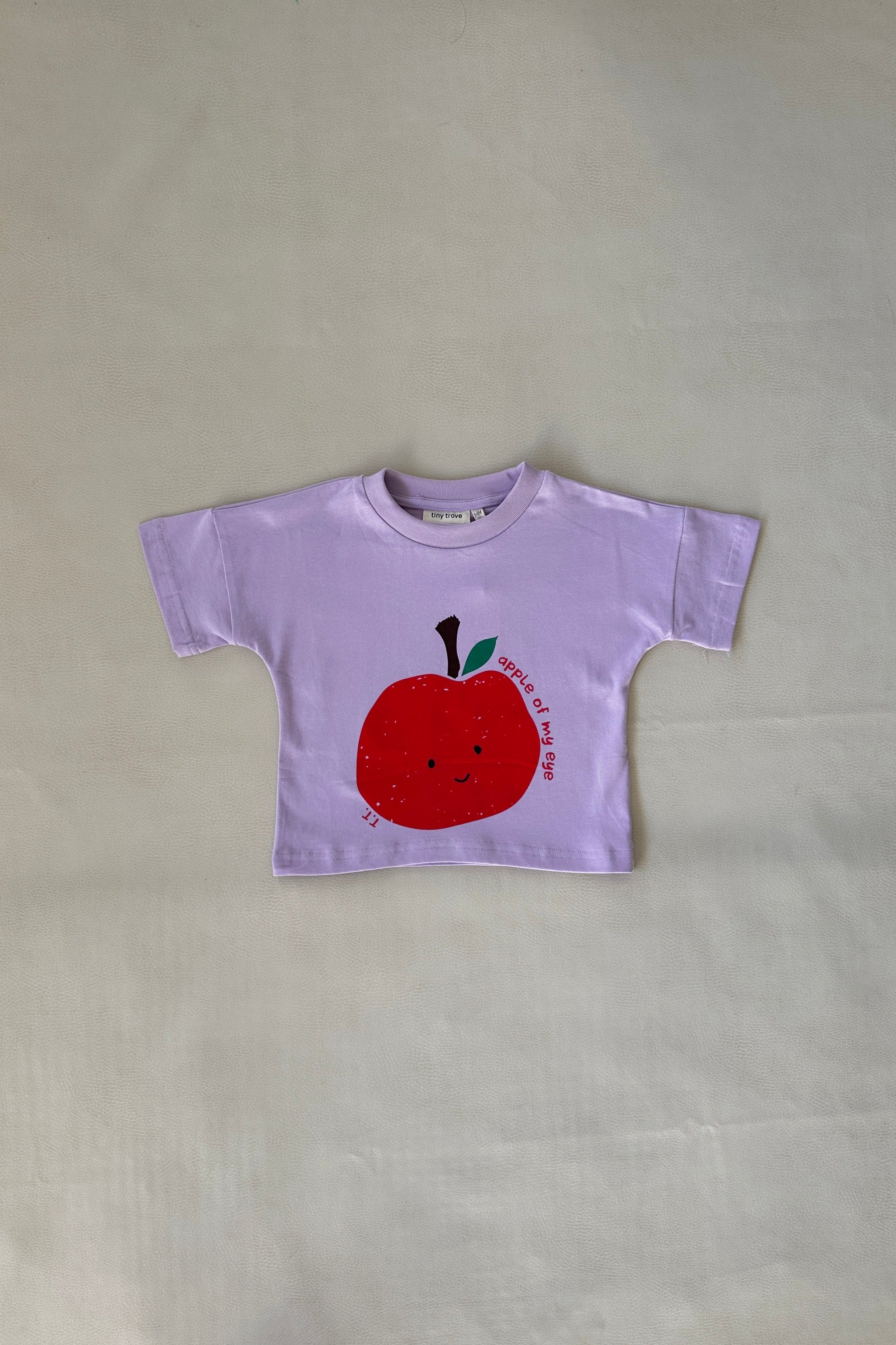 The Apple Relaxed Tee Lilac by TINY TROVE, size 6, showcases a cute smiling red apple graphic with "Sweet as apple pie." Made from soft cotton, this lavender-colored T-shirt offers a relaxed fit on a plain light backdrop.