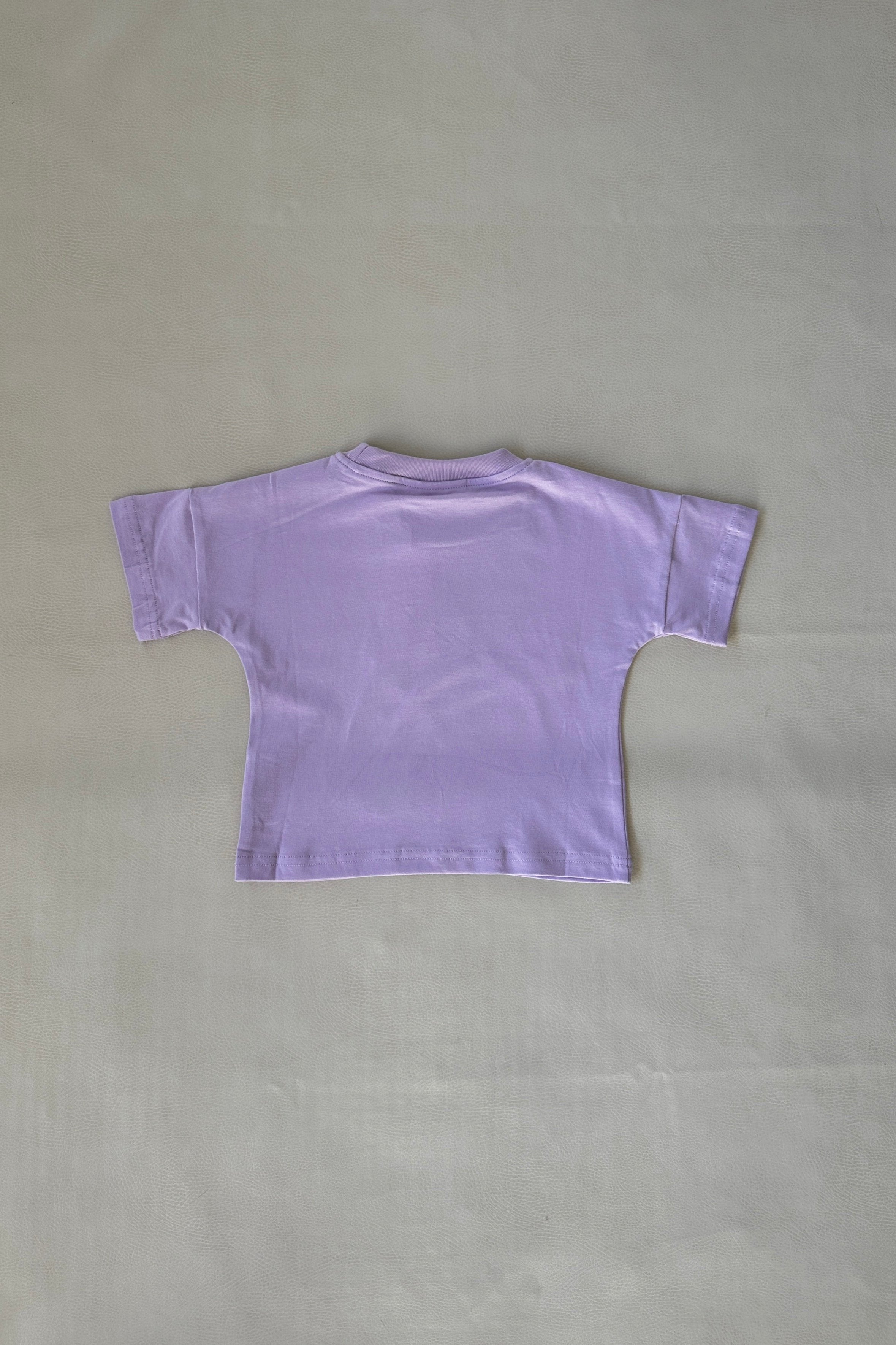 The Apple Relaxed Tee - Lilac by TINY TROVE is a straightforward, light purple, short-sleeved T-shirt, displayed flat on a plain off-white background. Made from 100% cotton, it features a relaxed fit and is shown from the back with a simple design devoid of logos or patterns.