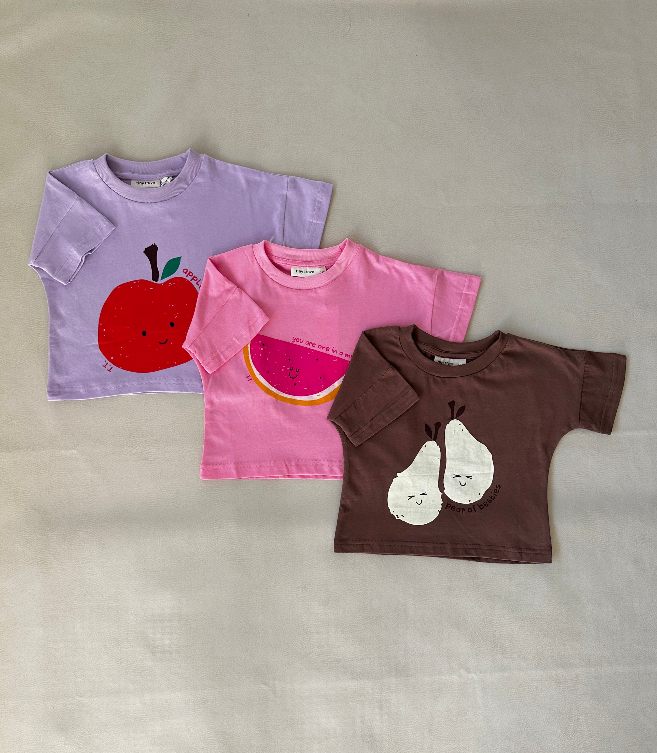 Three vibrant children's T-shirts from TINY TROVE are displayed on a flat surface. Made from cotton for a relaxed fit, the collection includes the Melon Relaxed Tee - Bubblegum in bright pink featuring a watermelon print, as well as a pastel purple shirt with an apple print and a brown tee adorned with pears, each exuding cheerful faces.