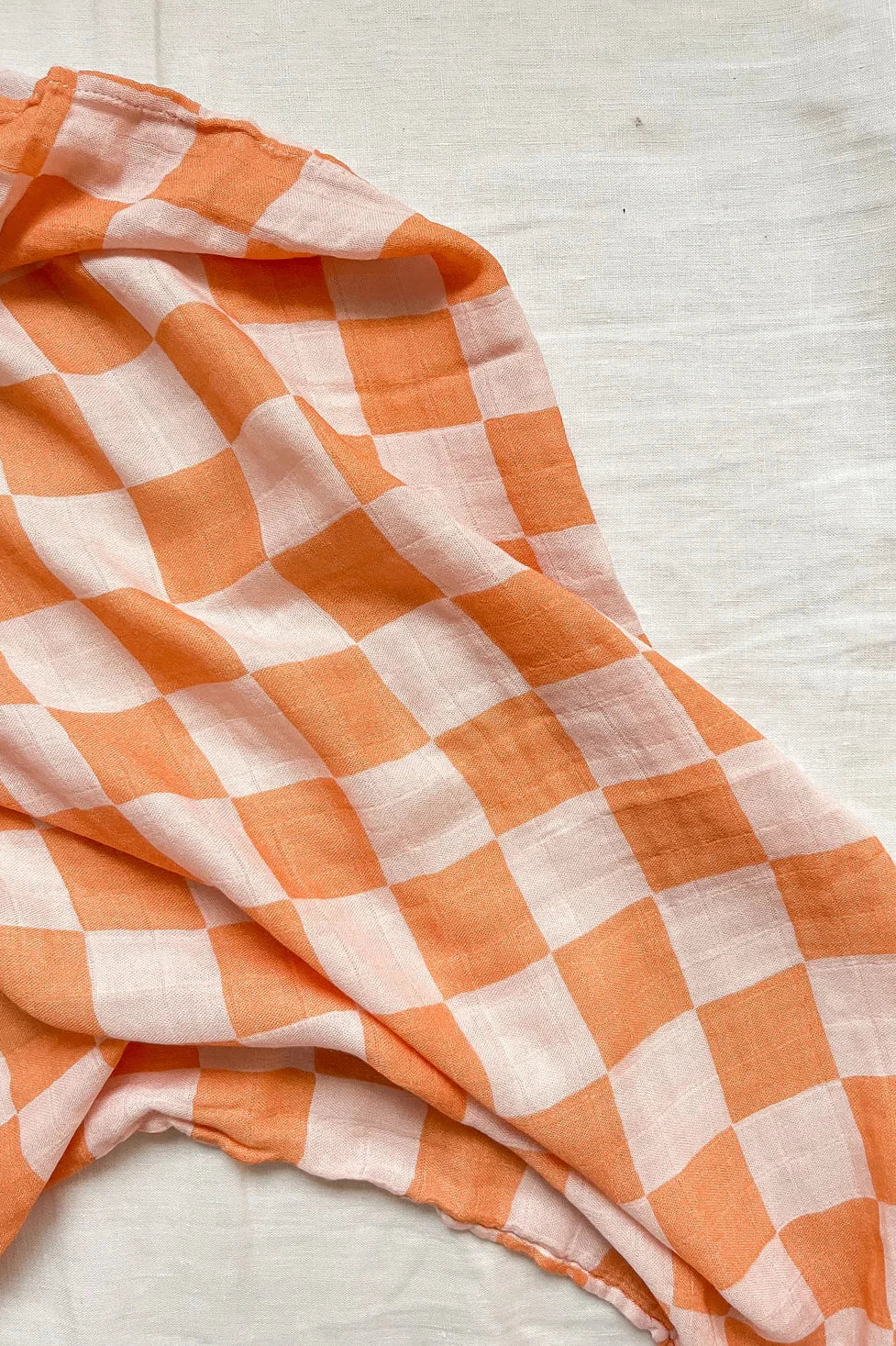 An Apricot Peaches Wrap, featuring MILKY DESIGNS' signature orange and white checkered pattern, is elegantly draped over a light-colored surface. The design consists of large, alternating squares, and the fabric appears to be lightweight with a hint of buttery softness.