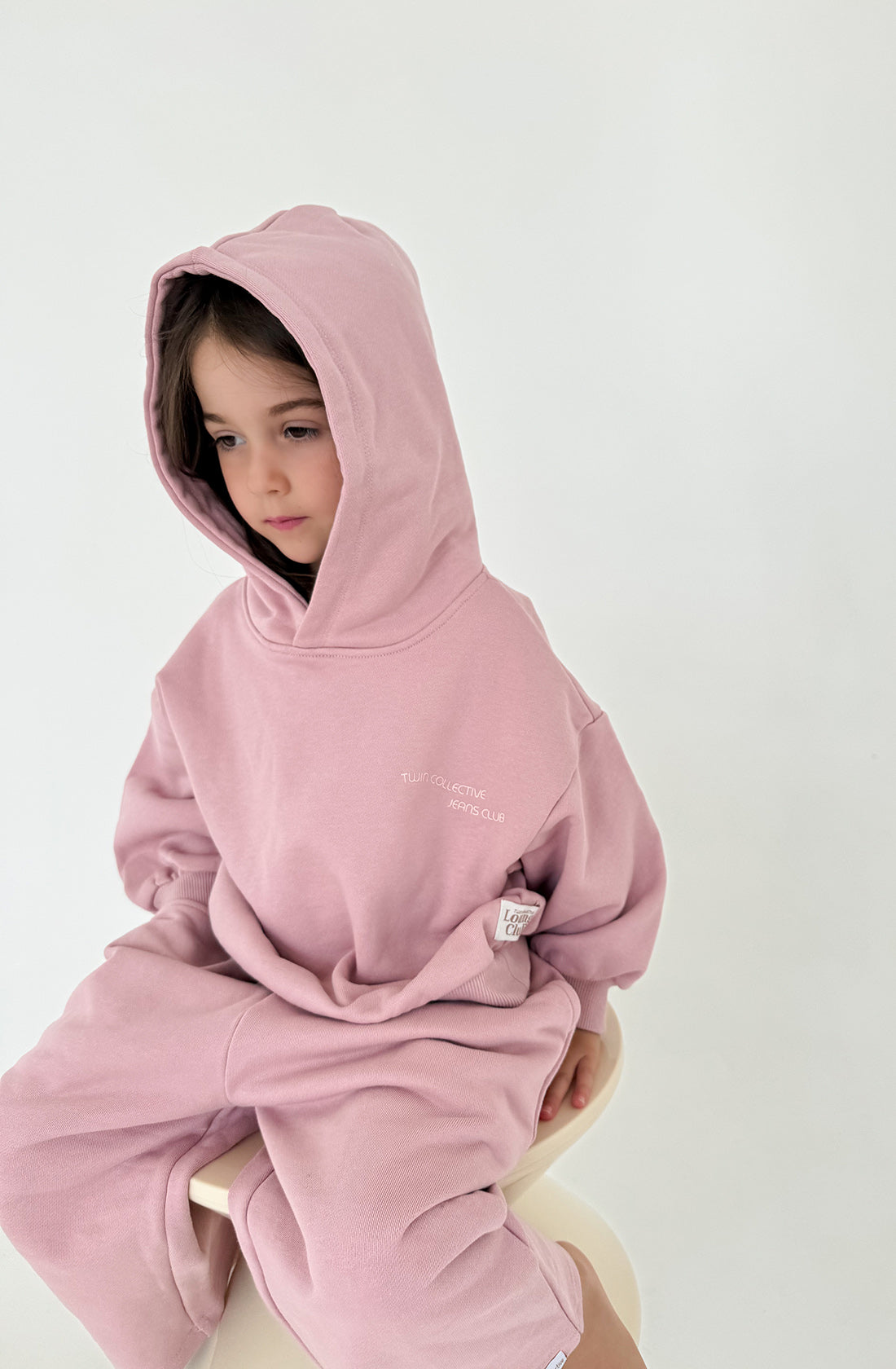 A young child wearing an oversized TWIN COLLECTIVE Logo Hoodie Set in Pink Haze, featuring a heavy loopback terry fleece hoodie and matching oversized track pants, sits on a round stool. The background is a plain white wall, giving the image a minimalist aesthetic. The child appears to be looking down with a calm expression.