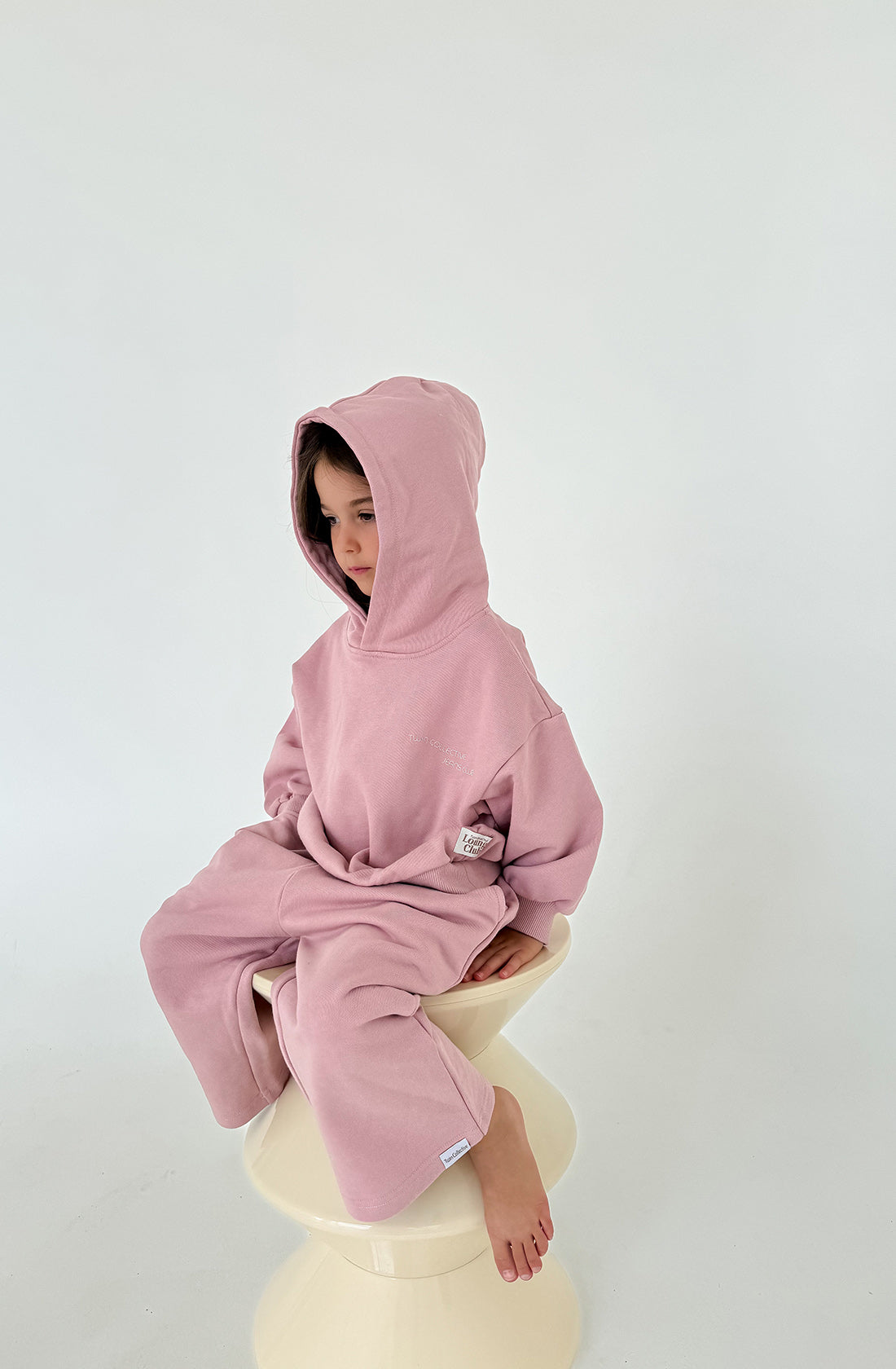 A young child, dressed in an oversized Logo Hoodie Set Pink Haze by TWIN COLLECTIVE, sits on a white chair. The child's pastel pink hoodie and matching track pants, both crafted from heavy loopback terry fleece, create a cozy ensemble. With the hood up partially covering their face, the child gazes downward against a plain white backdrop for a minimalist effect.