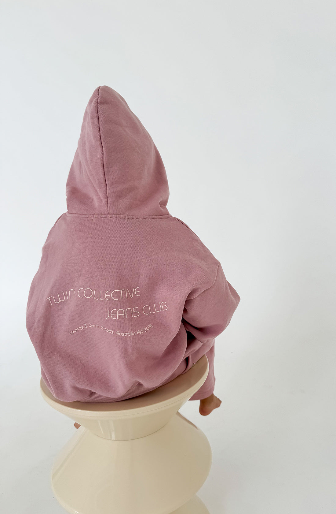 A person in a light pink Logo Hoodie Set Pink Haze by TWIN COLLECTIVE, made of heavy loopback terry fleece with the text "Twin Collective Jeans Club" on the back, sits on a beige stool. They are facing away from the camera, with the hood up over their head. The background is plain and white.