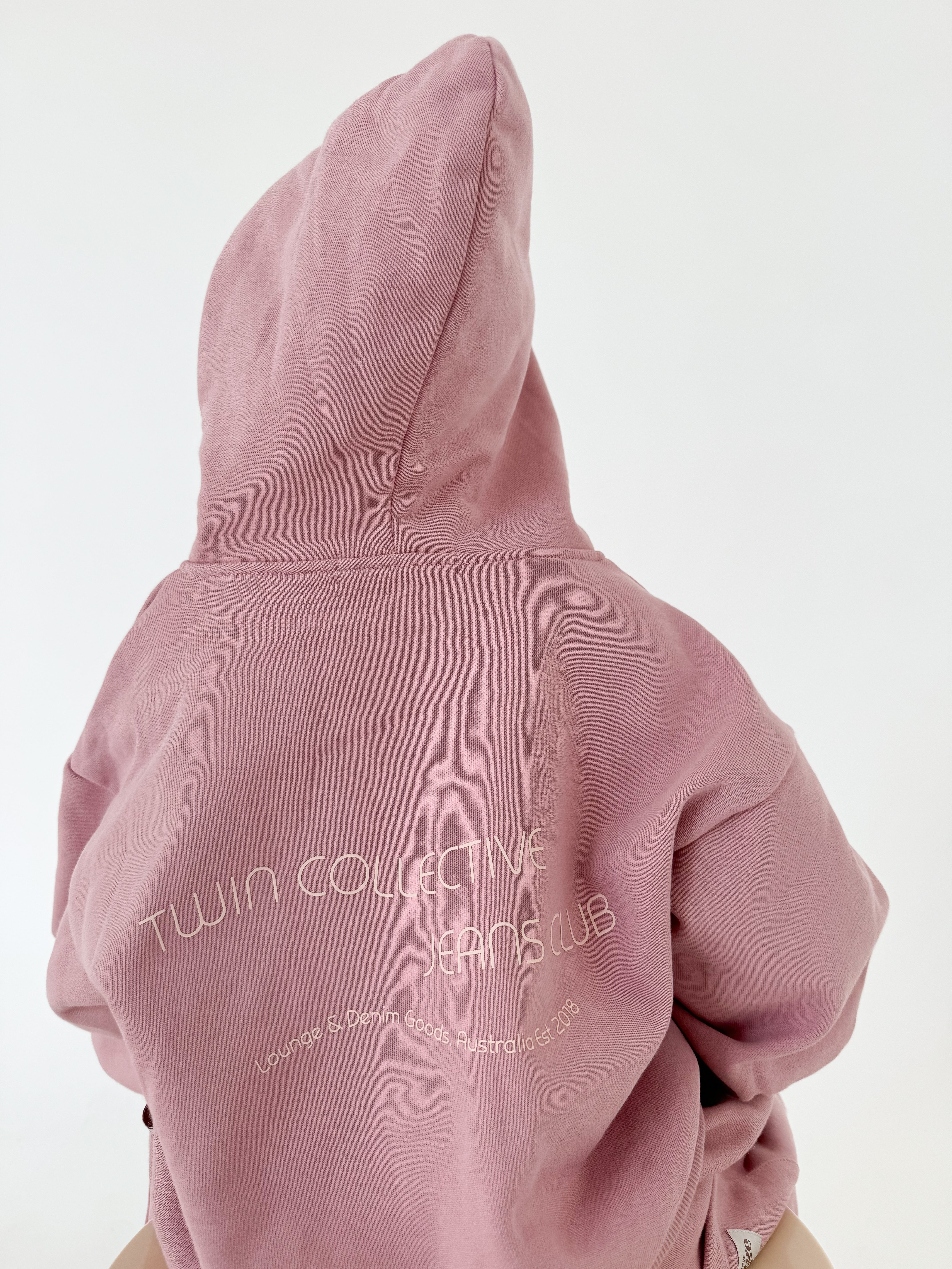 A person wearing a pink "Logo Hoodie Set Pink Haze" with the hood up, seen from the back. The TWIN COLLECTIVE hoodie showcases "TWIN COLLECTIVE JEANS CLUB" in white text and "Lounge & Denim Goods, Australia Est. 2013" beneath in a smaller font. They're likely pairing it with oversized track pants for ultimate comfort.