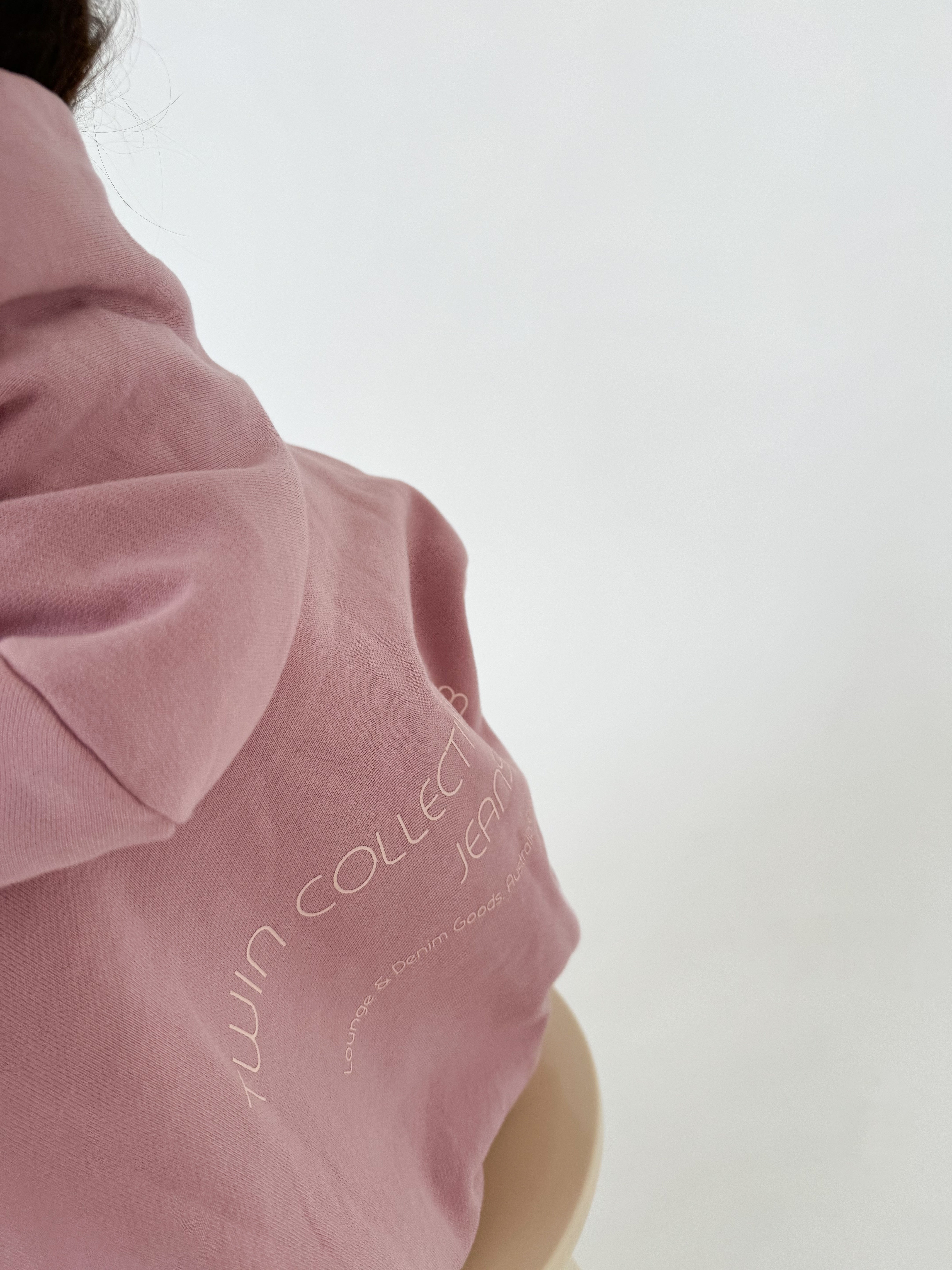 Close-up of the back of a person wearing a pink TWIN COLLECTIVE Logo Hoodie Set Pink Haze made from heavy loopback terry fleece, with the words "TWIN COLLECTIVE" and partially visible text printed in white. The plain, neutral background highlights the fabric and text on the hoodie.