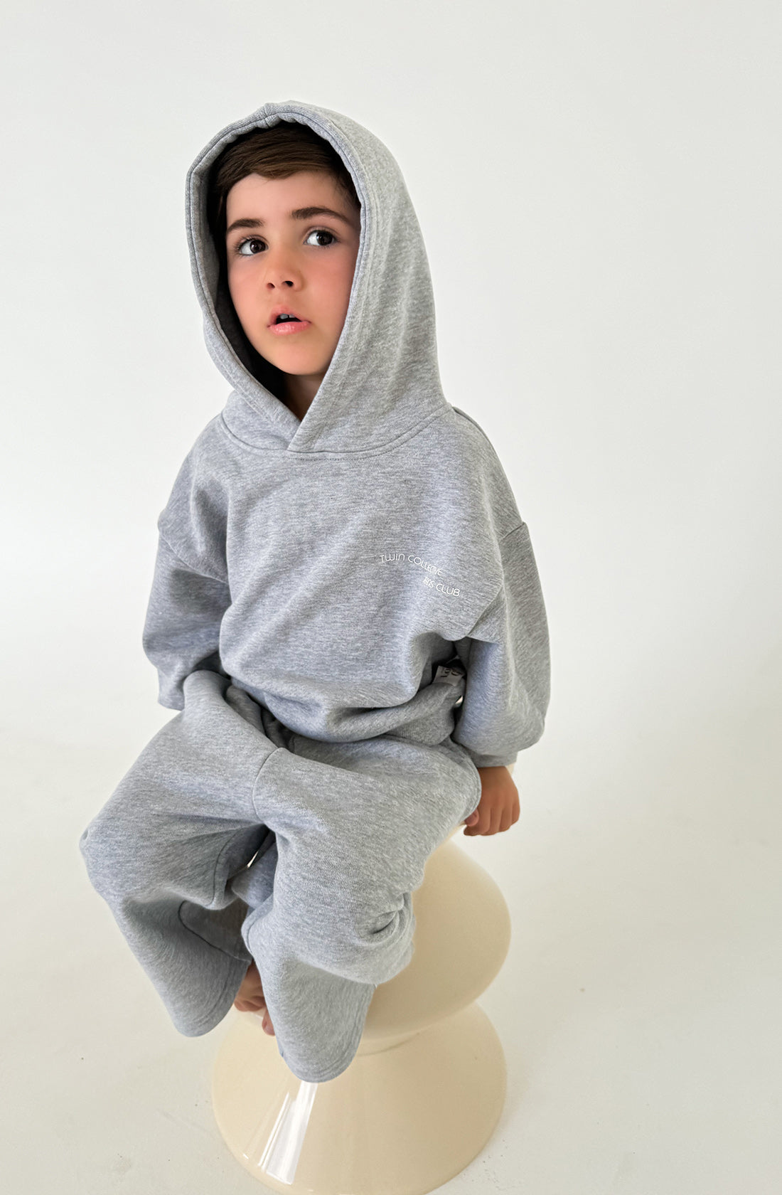 A young child with dark hair is wearing a grey Marle Logo Hoodie Set from TWIN COLLECTIVE, made from Loopback Terry fleece. The child is sitting on an off-white stool against a plain, light-colored background, looking slightly to the side with a relaxed expression.