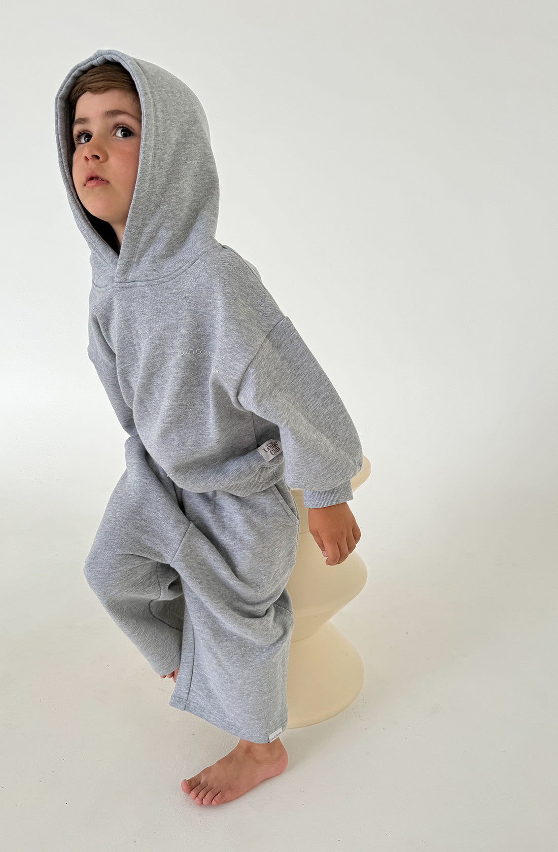 A young child wearing a TWIN COLLECTIVE Logo Hoodie Set Grey Marle, featuring a loose-fitting Lounge Club Hoodie in gray Loopback Terry fleece and matching oversized track pants, stands on a modern, unconventional stool in a minimalistic room with a white background. The child looks slightly upward and to the side with a thoughtful expression.