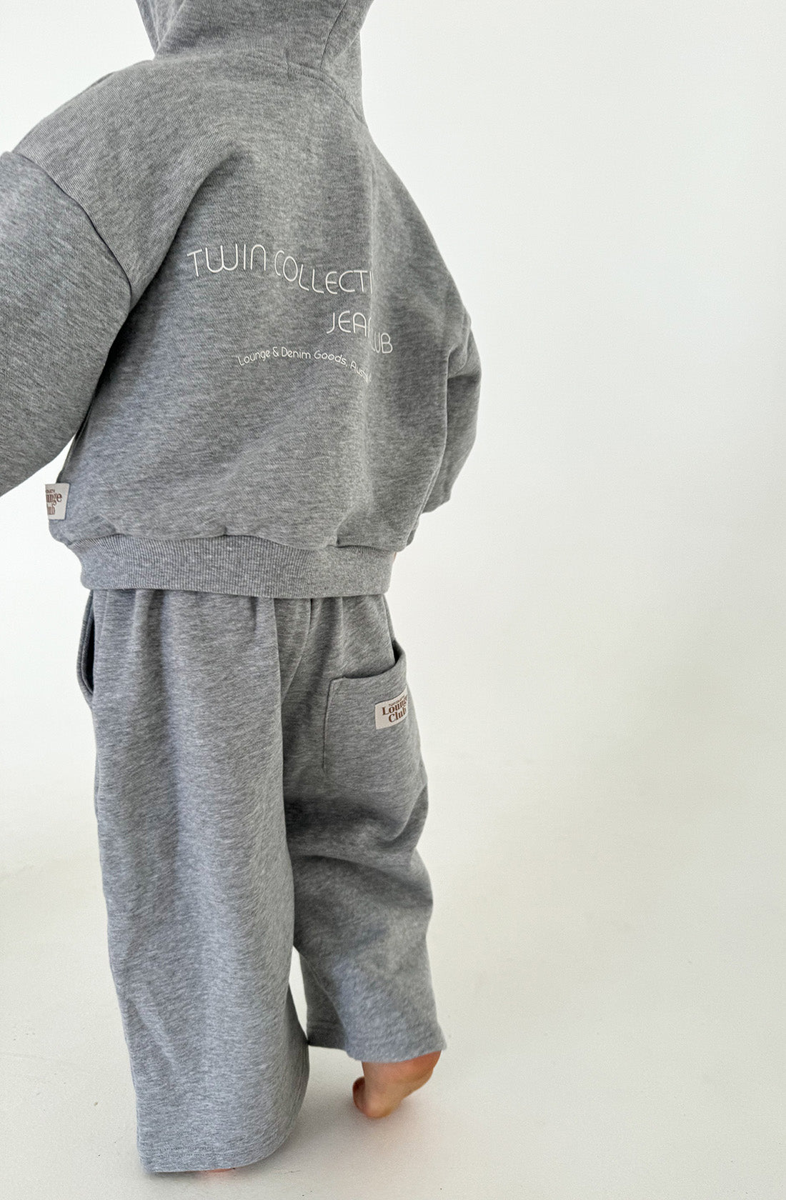 A child dressed in a Logo Hoodie Set Grey Marle stands facing away. The back of the Lounge Club Hoodie reads "TWIN COLLECTIVE JEAN CLUB" in white text. The outfit, made from soft Loopback Terry fleece, boasts a casual, comfortable style against a plain white background.