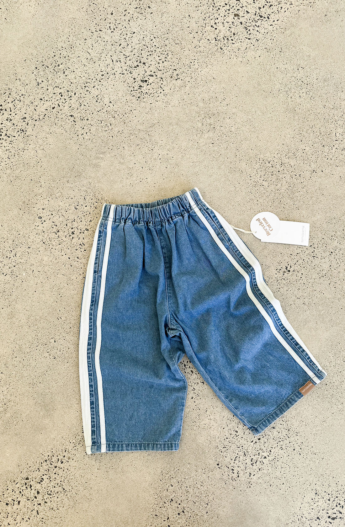 A pair of Tricky Track Pant blue denim shorts with white side stripes lies on a speckled concrete surface, showcasing a vintage-inspired design. These TWIN COLLECTIVE shorts feature an elastic waistband and a small brown tag on one leg. A white tag with branding is attached to the waistband by a plastic fastener.